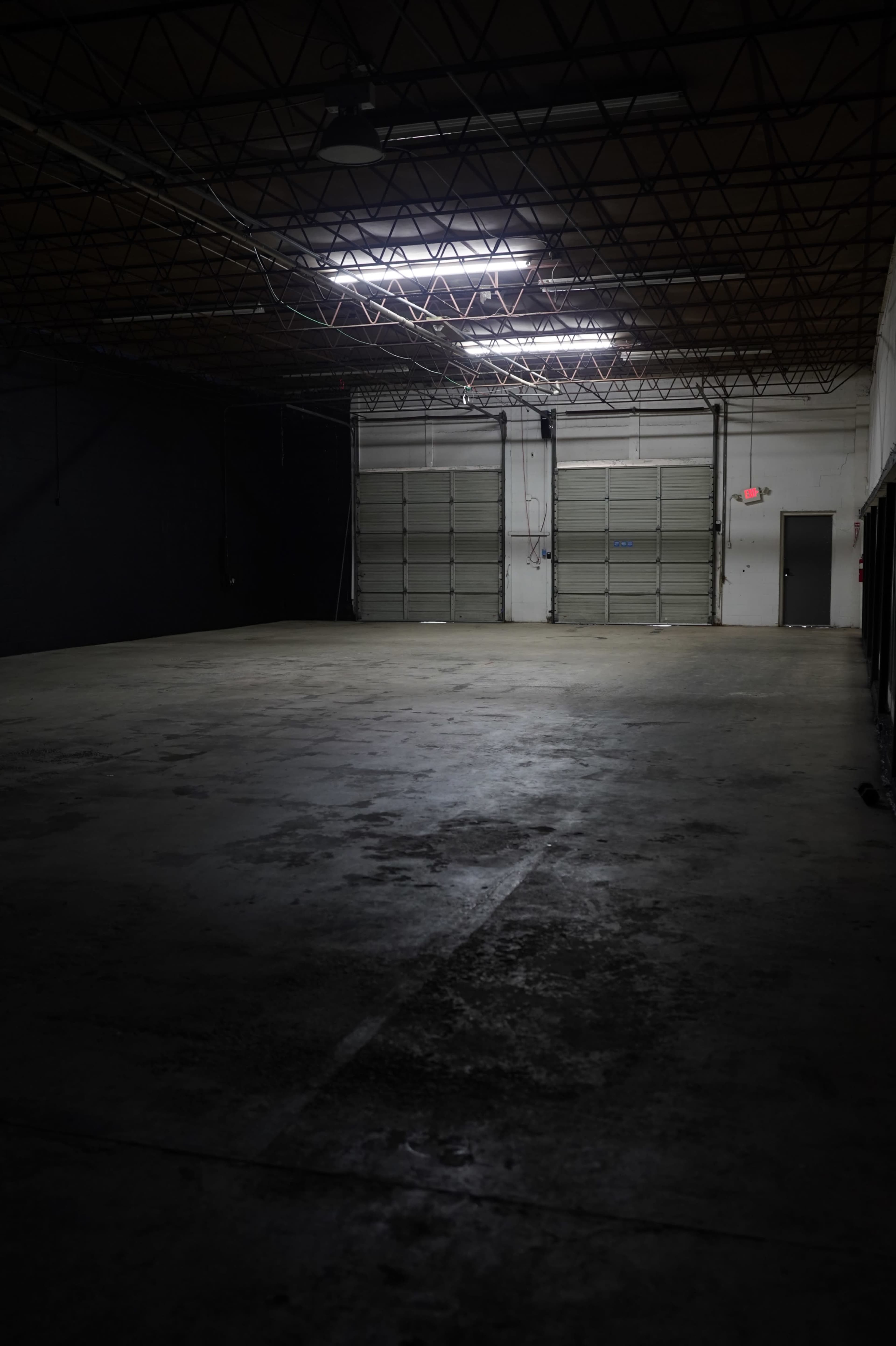 The image shows a dimly lit, empty industrial warehouse with a concrete floor and large roll-up doors.