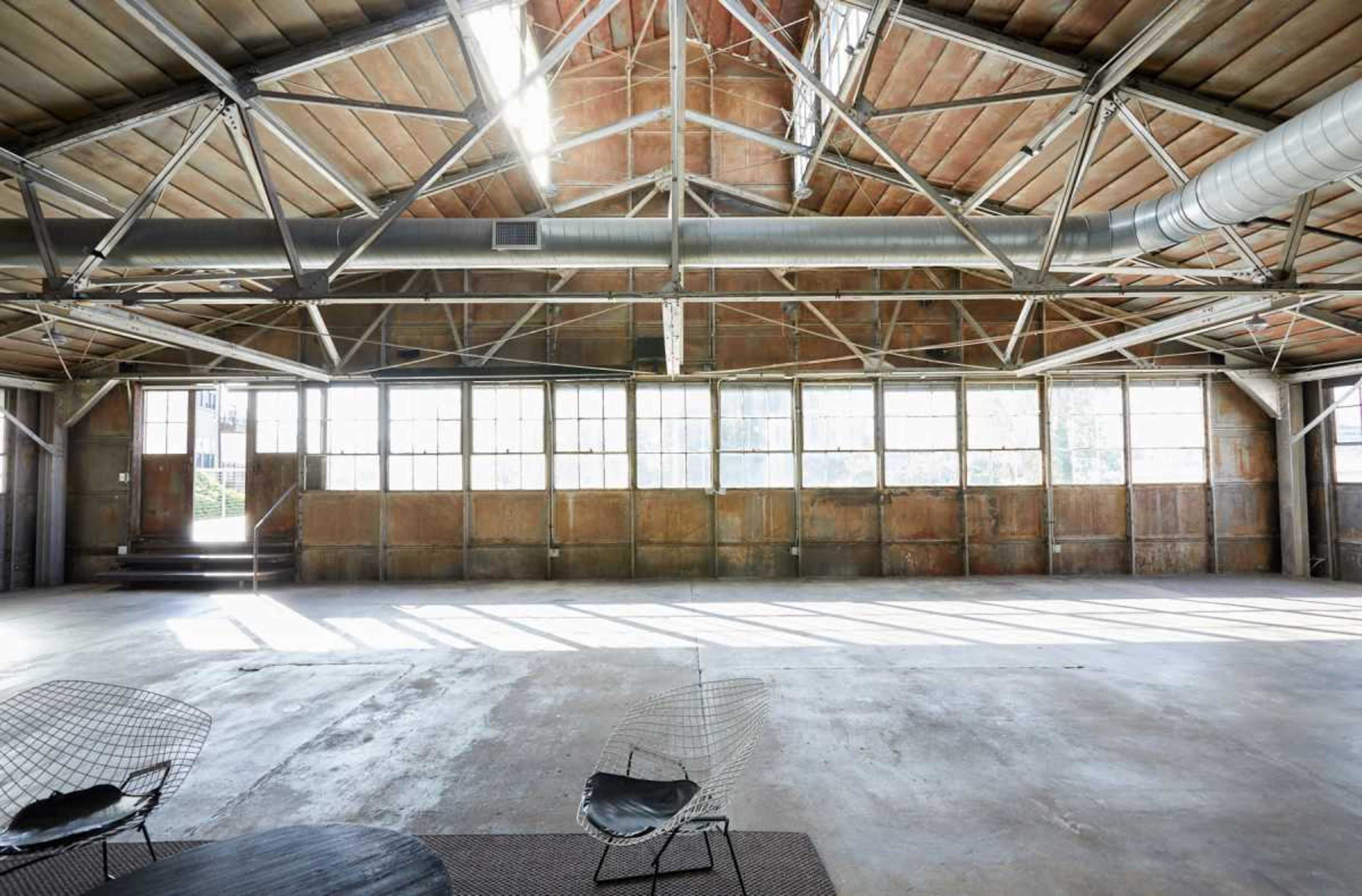 The image shows a spacious industrial-style interior with high ceilings, large windows allowing natural light, and minimal furnishings.