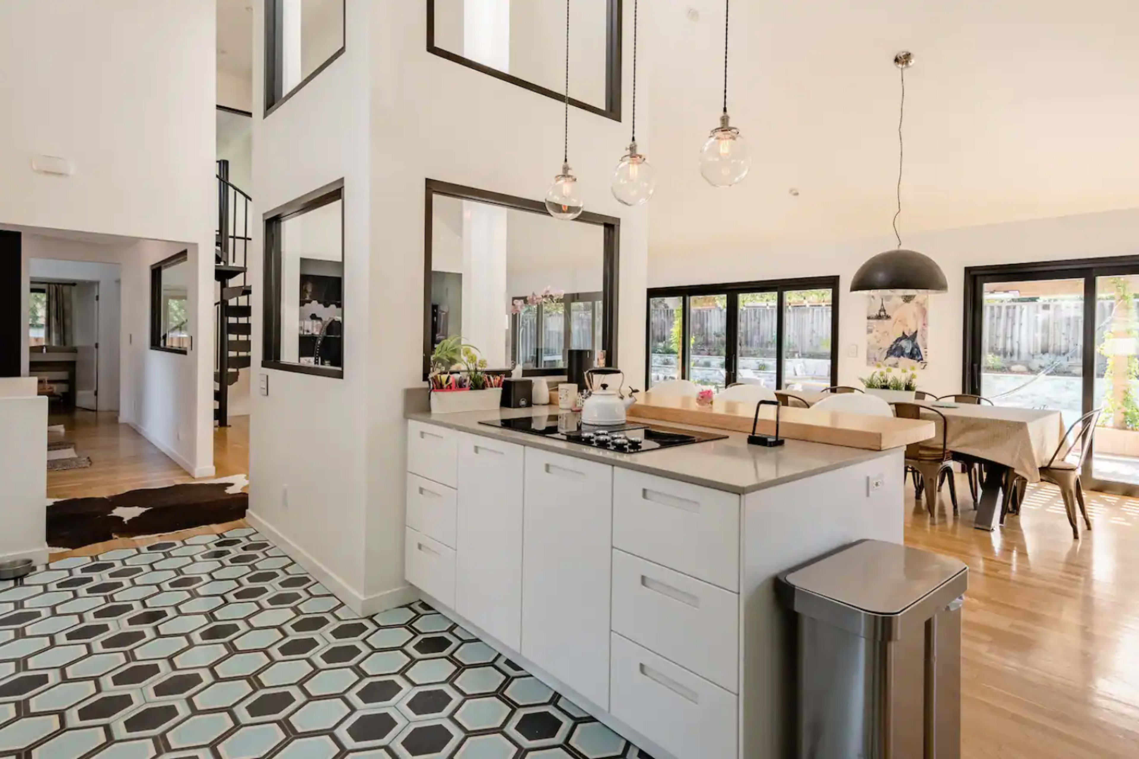 The image shows a modern kitchen with white cabinetry, a central island, and a patterned tile floor, adjacent to a dining area with large windows.