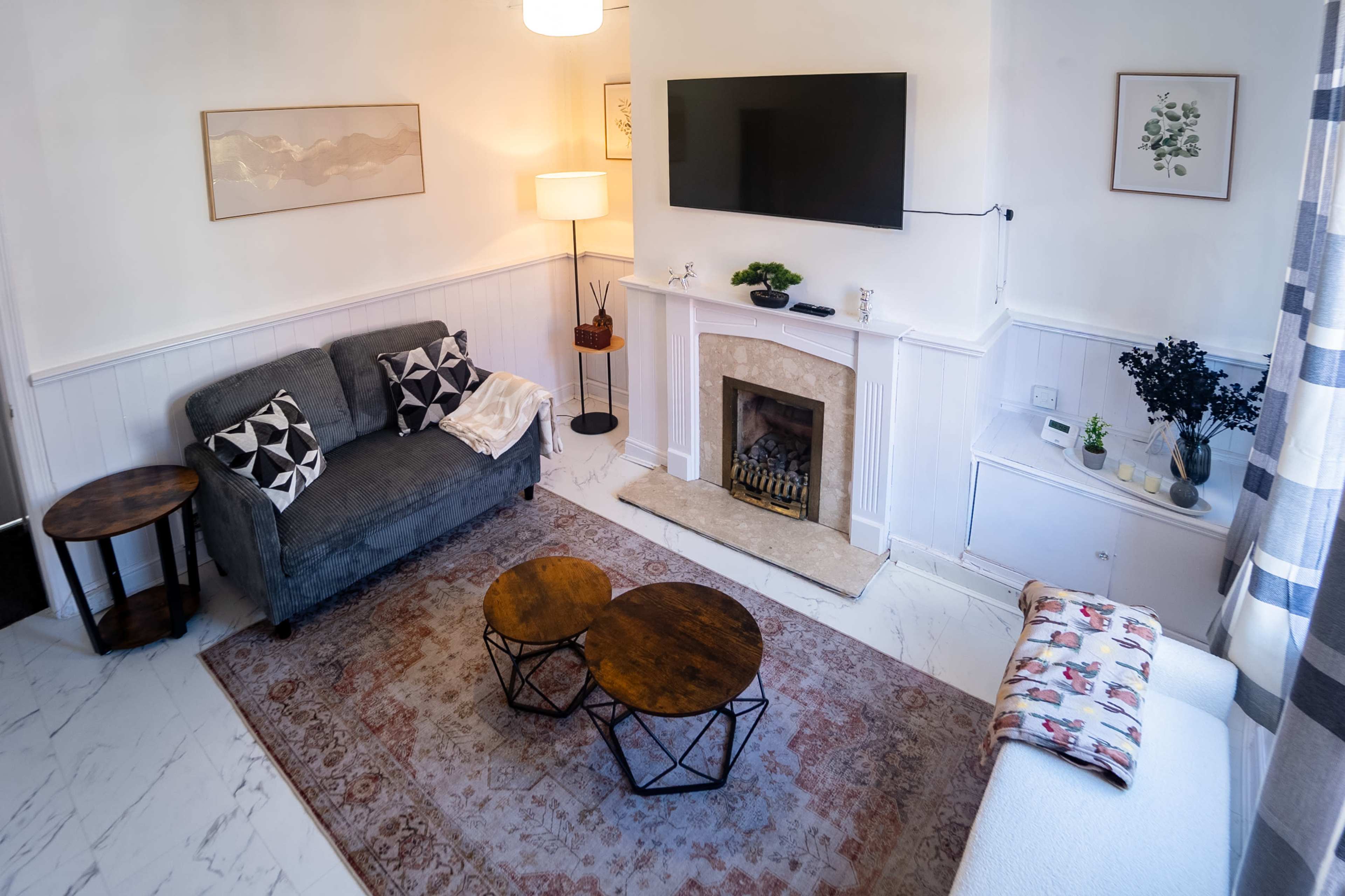 The image shows a cozy living room featuring a gray sofa, a fireplace with a television mounted above it, two round wooden coffee tables, and a rug on the floor.