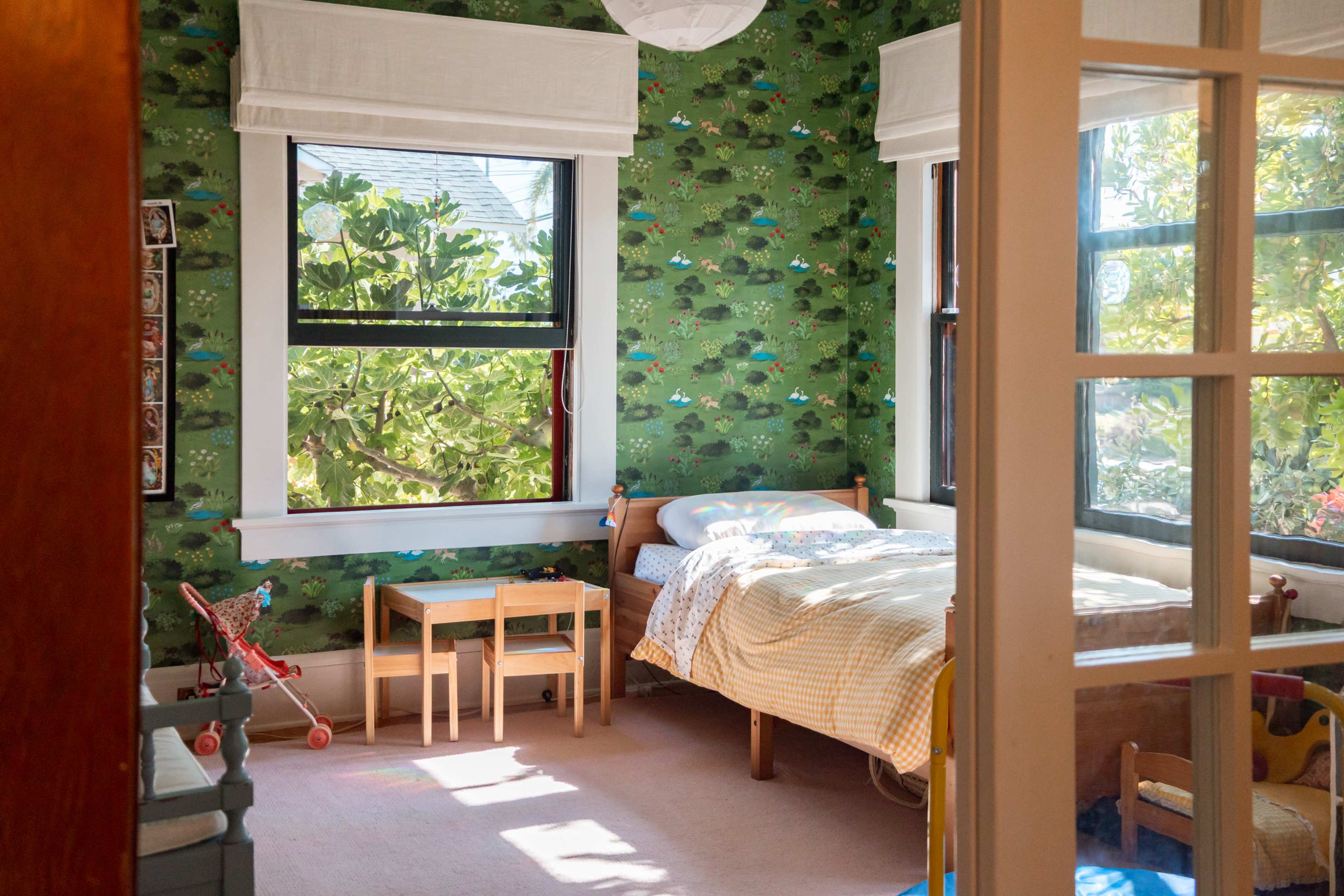 The image shows a sunlit children's bedroom with patterned green wallpaper, a bed with a colorful blanket, and a small table set beside the bed.