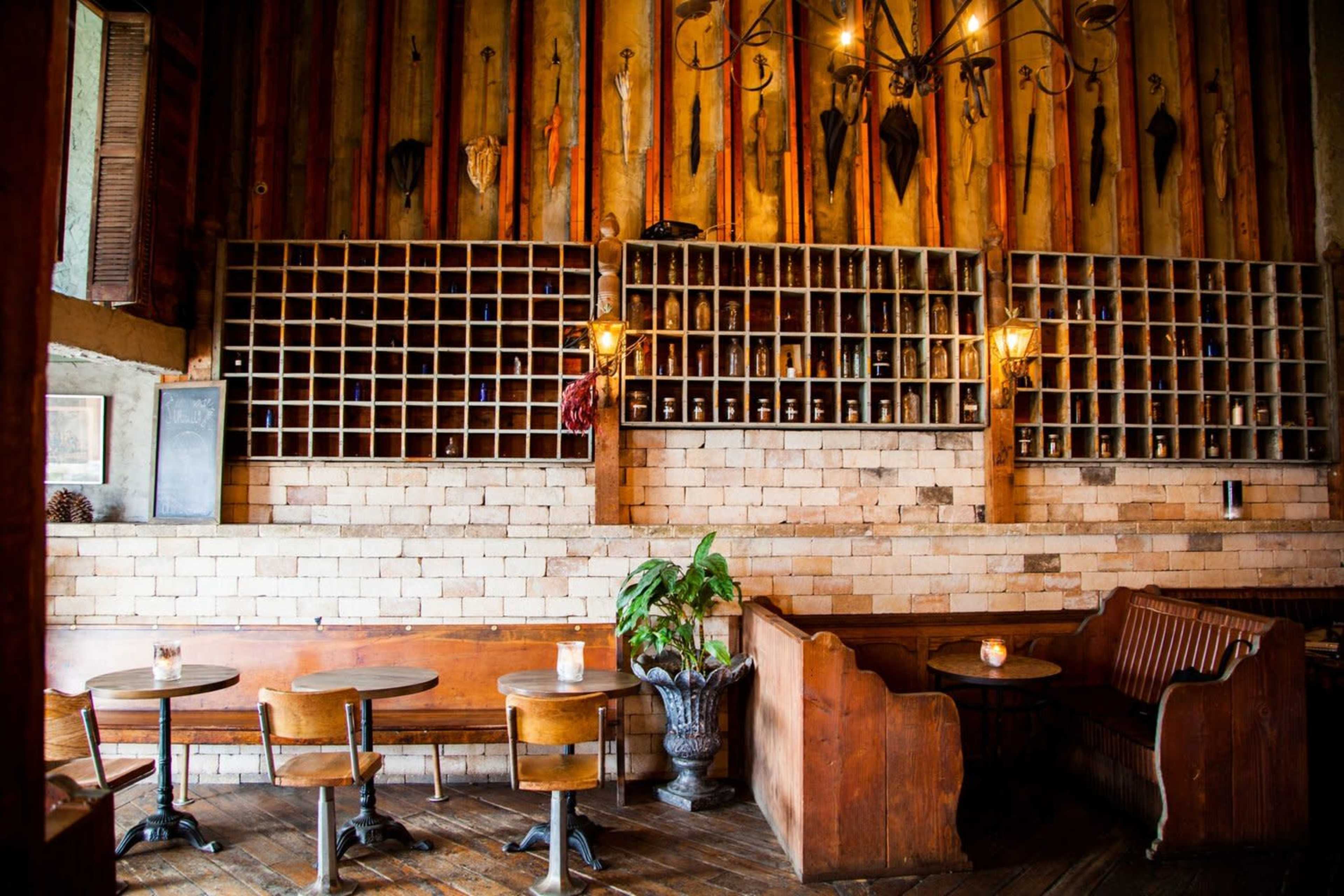 A rustic restaurant features wooden tables and chairs, a wall of shelves displaying bottles, and a decorative arrangement of umbrellas hanging above.