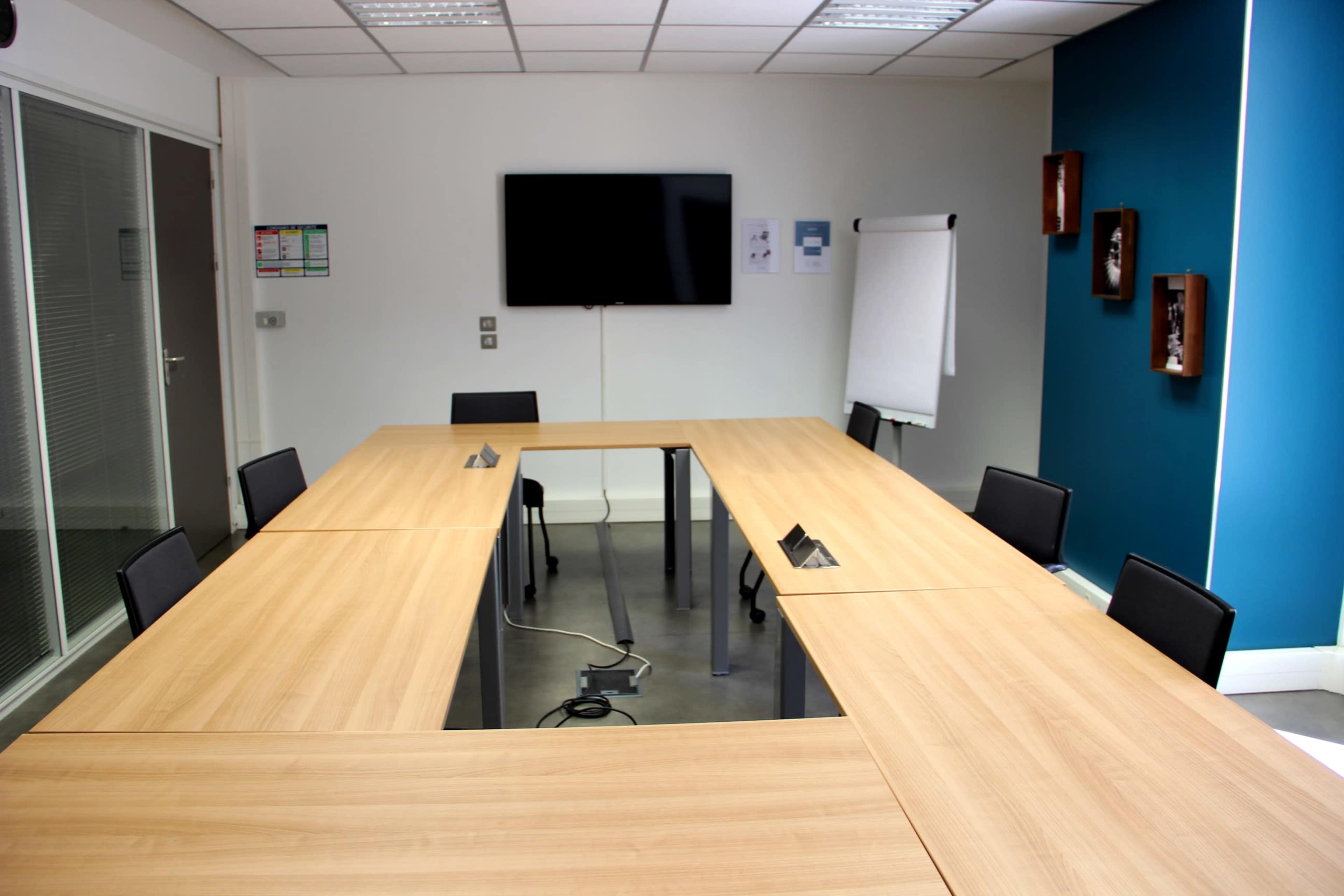 A modern conference room features a long wooden table, several black chairs, a wall-mounted television, and a whiteboard in a bright professional setting.