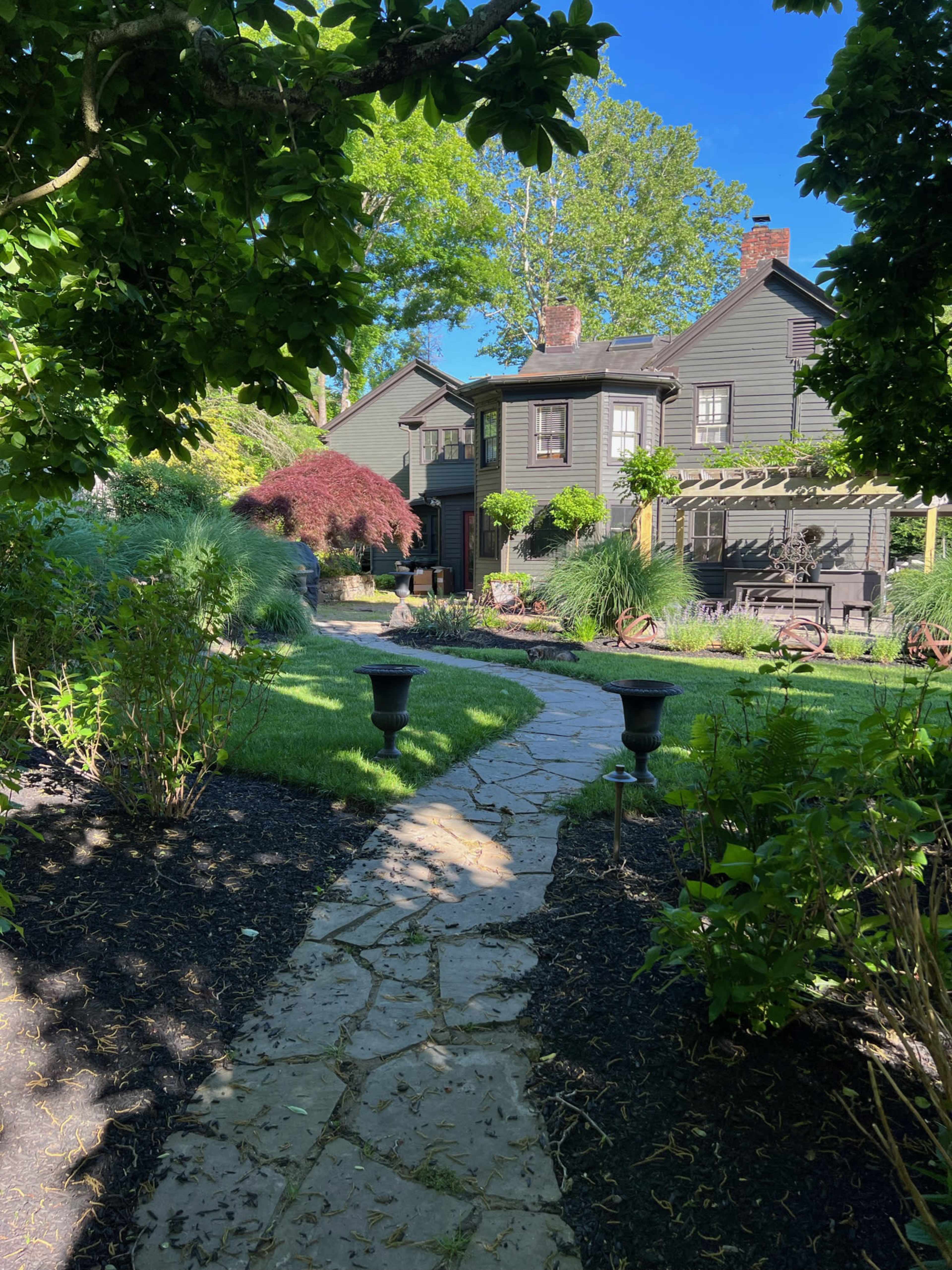 A stone path leads through a landscaped garden to a dark-colored house with multiple stories and a porch.