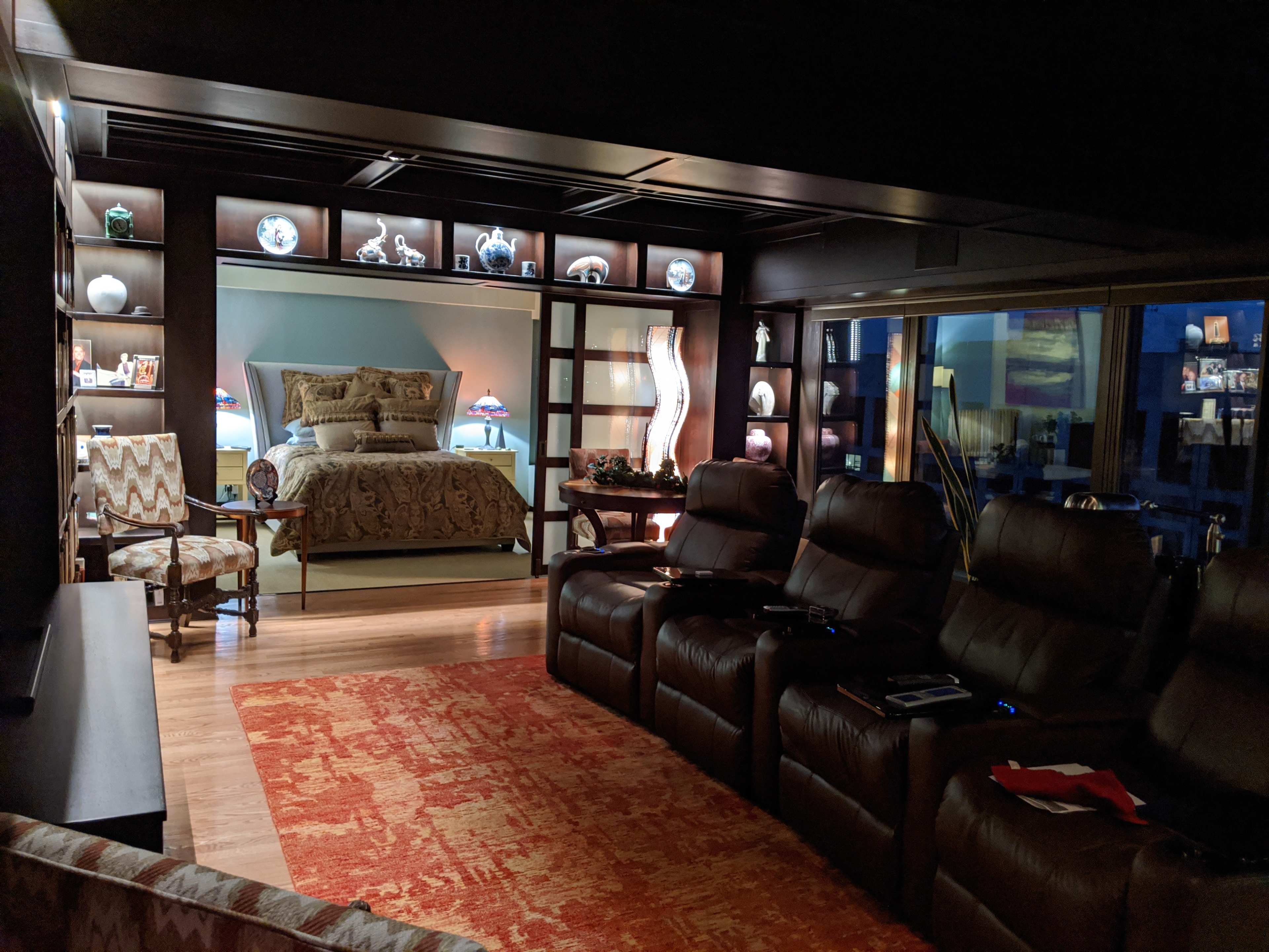 A cozy, dimly lit room featuring a bed with decorative pillows, flanked by shelves displaying various items, and a seating area with reclining chairs in front of a television.