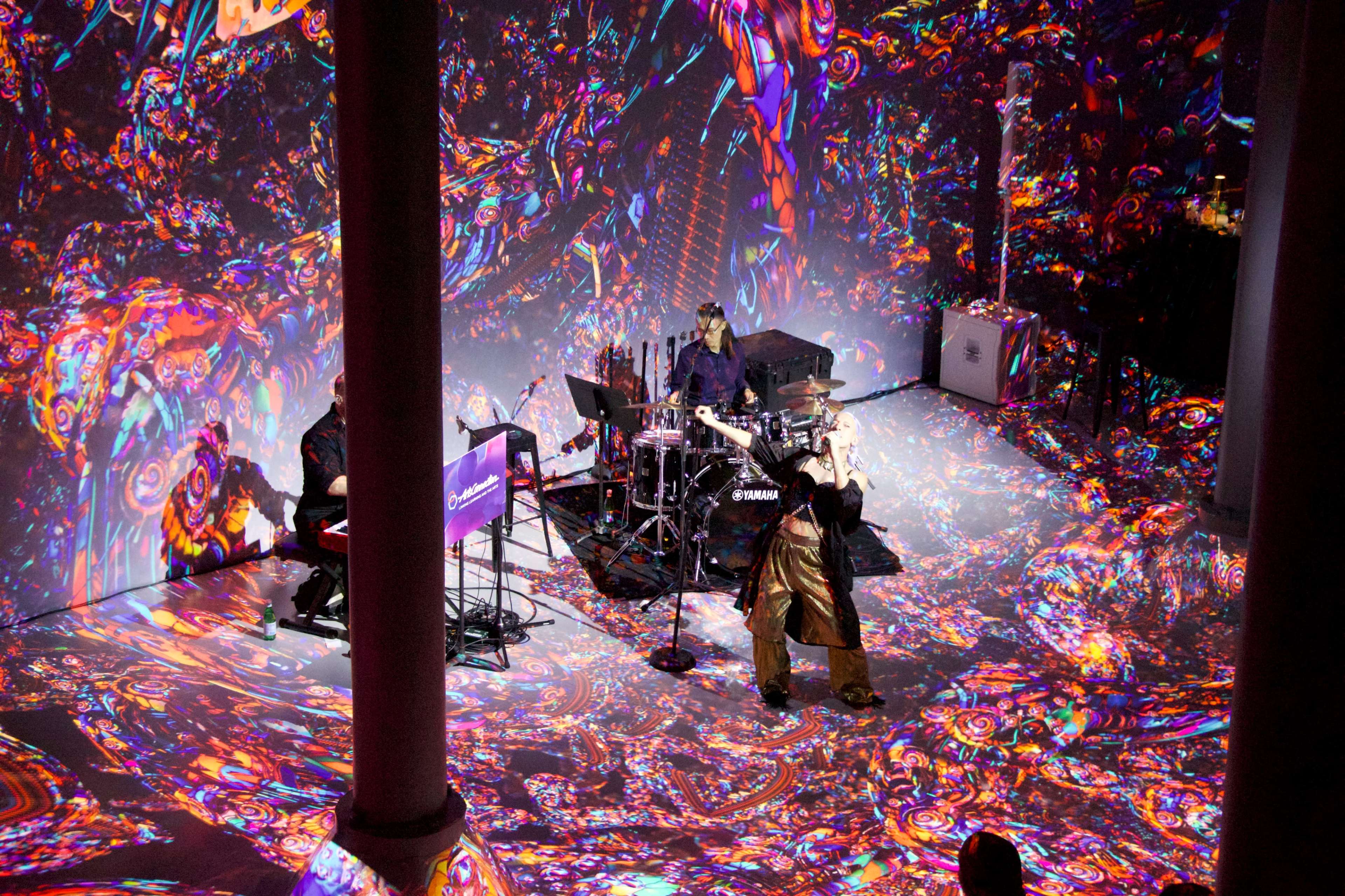 A musician performs on stage surrounded by vibrant, colorful projections that extend across the walls and floor of the venue.