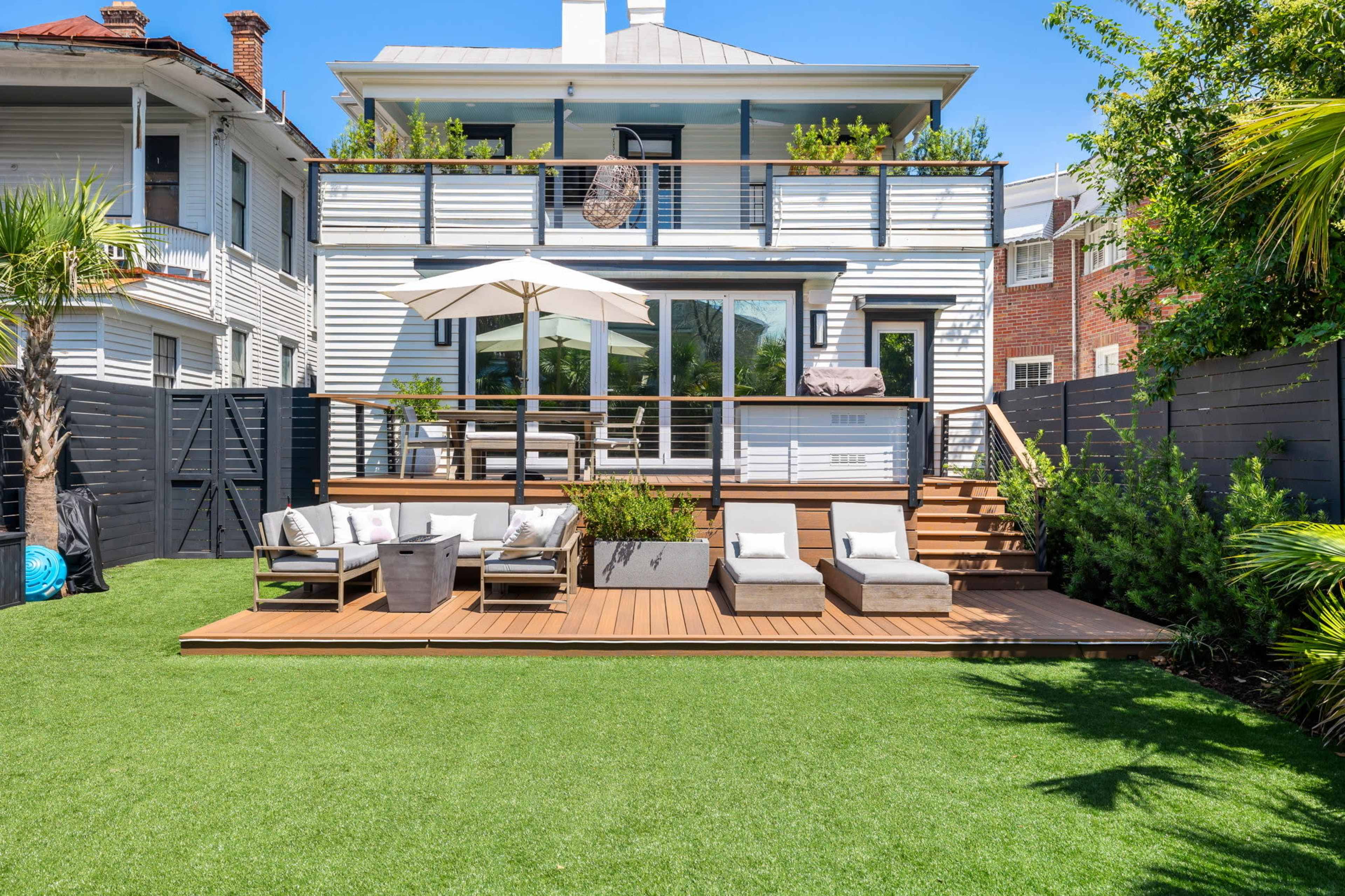 The image shows a modern backyard patio with a wooden deck, lounge chairs, and an umbrella, surrounded by greenery and residential buildings.
