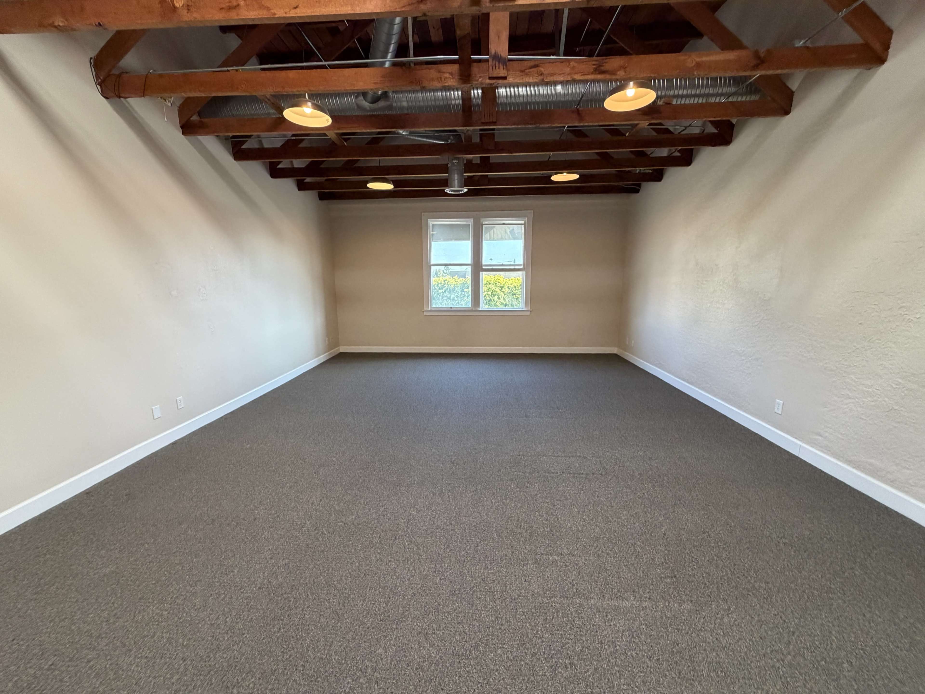 The image shows an empty room with a carpeted floor, exposed wooden beams on the ceiling, and a window allowing natural light to enter.