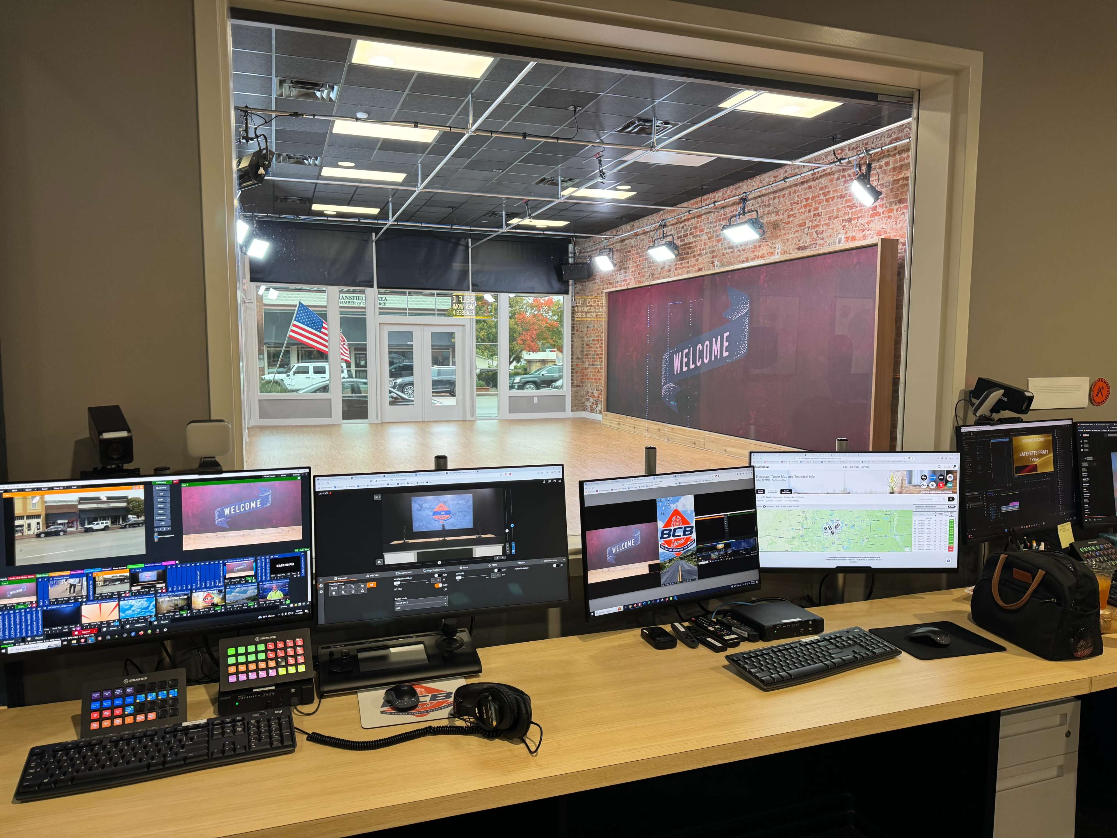 A control room with multiple computer monitors and equipment overlooking a studio space featuring a "Welcome" sign and an American flag.