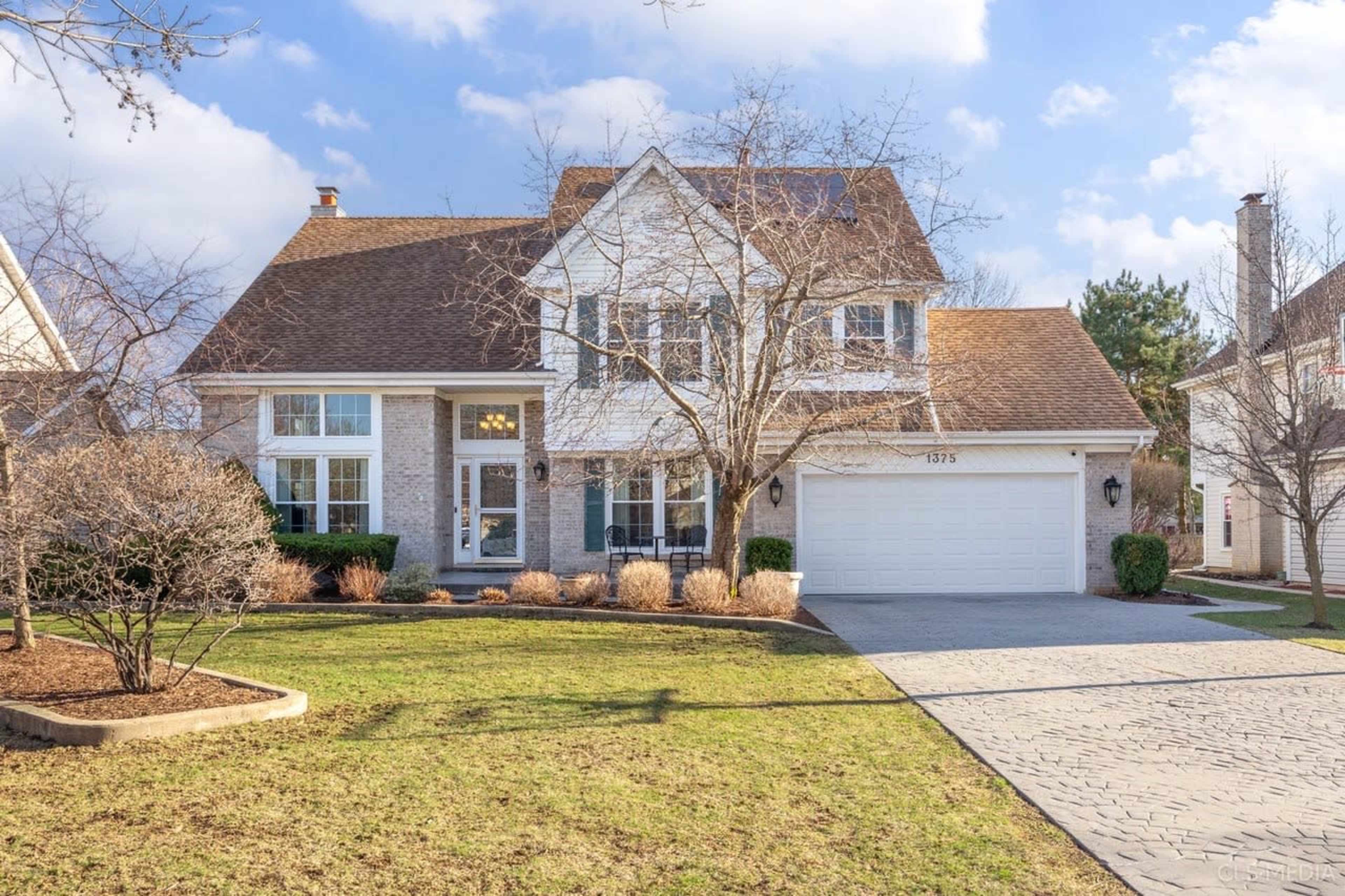 The image shows a two-story house with a brown shingle roof, brick facade, and a landscaped front yard.