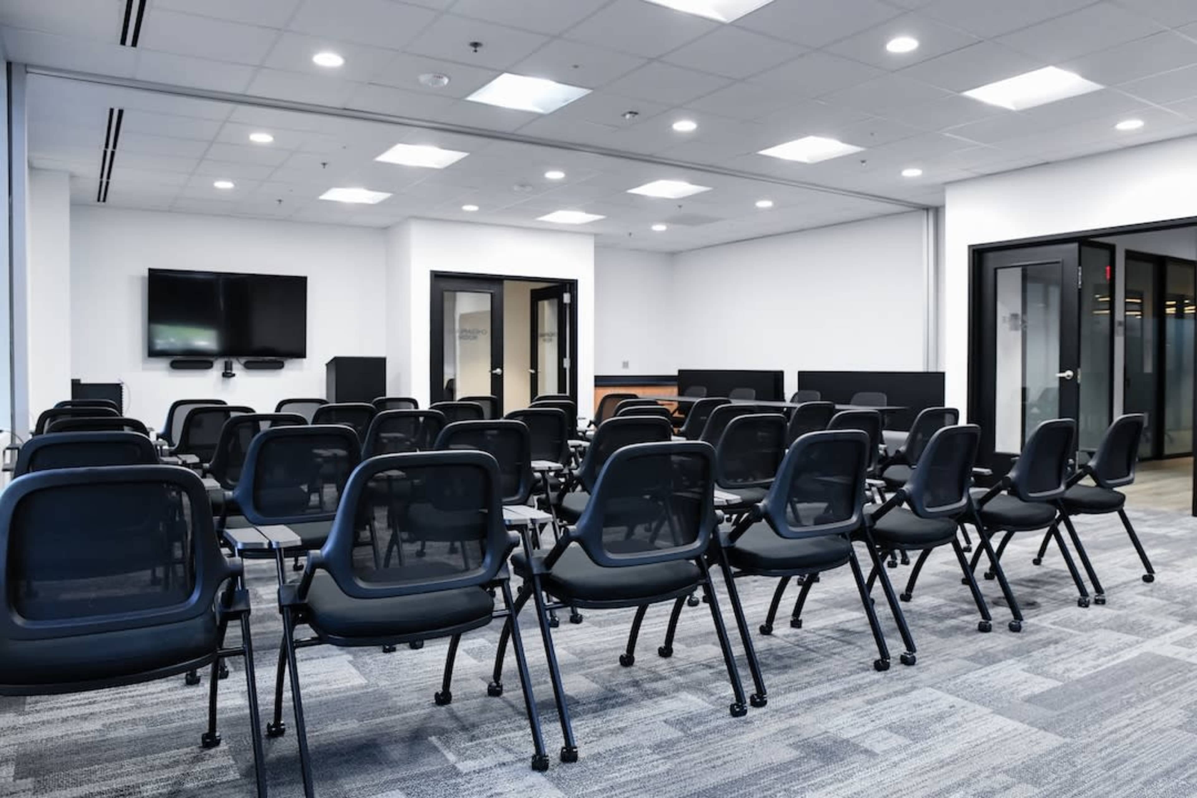 A modern meeting room features rows of black chairs facing a wall-mounted screen.