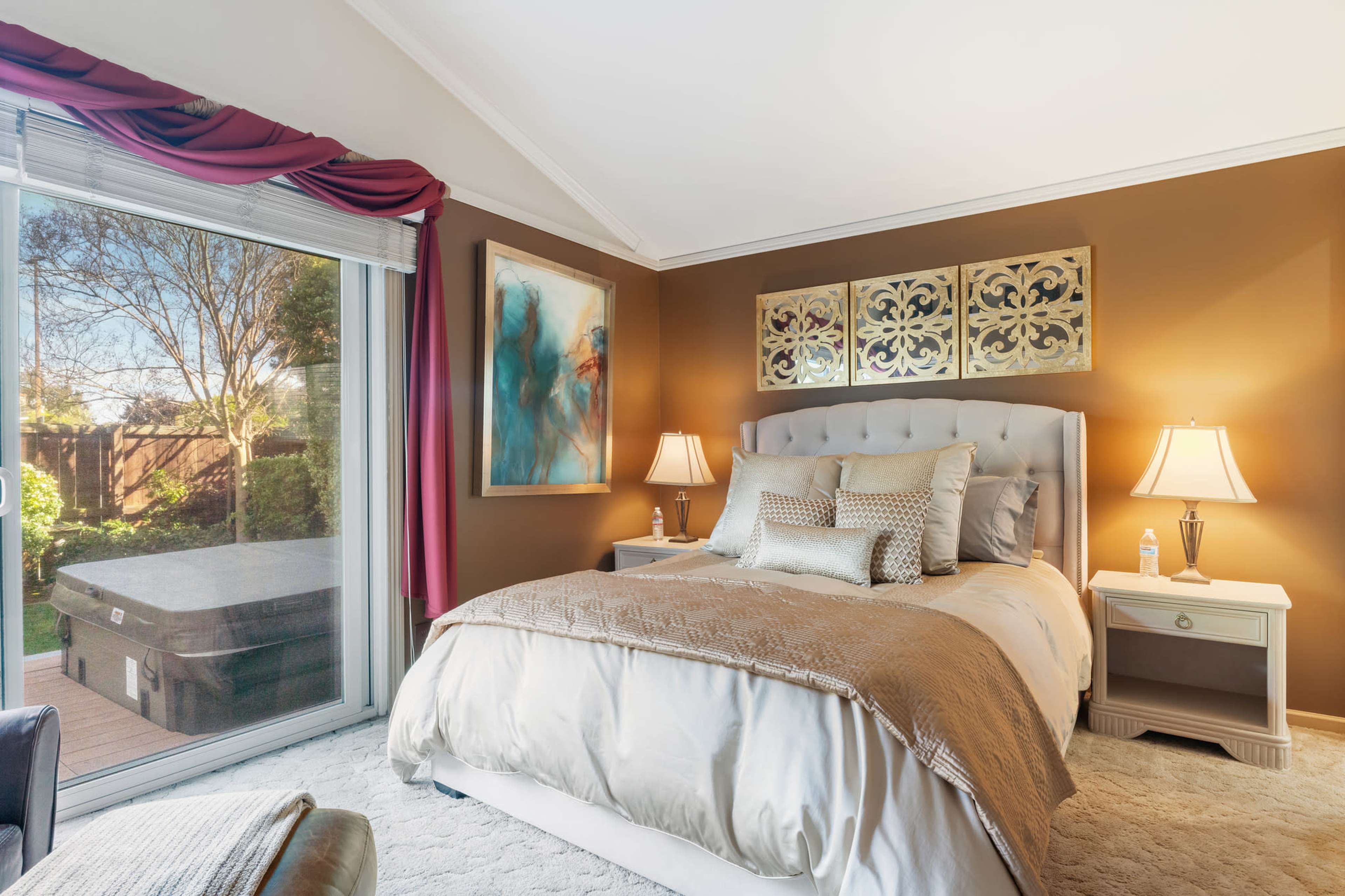A well-decorated bedroom featuring a large bed, bedside tables with lamps, and sliding glass doors that lead to an outdoor area with a hot tub.