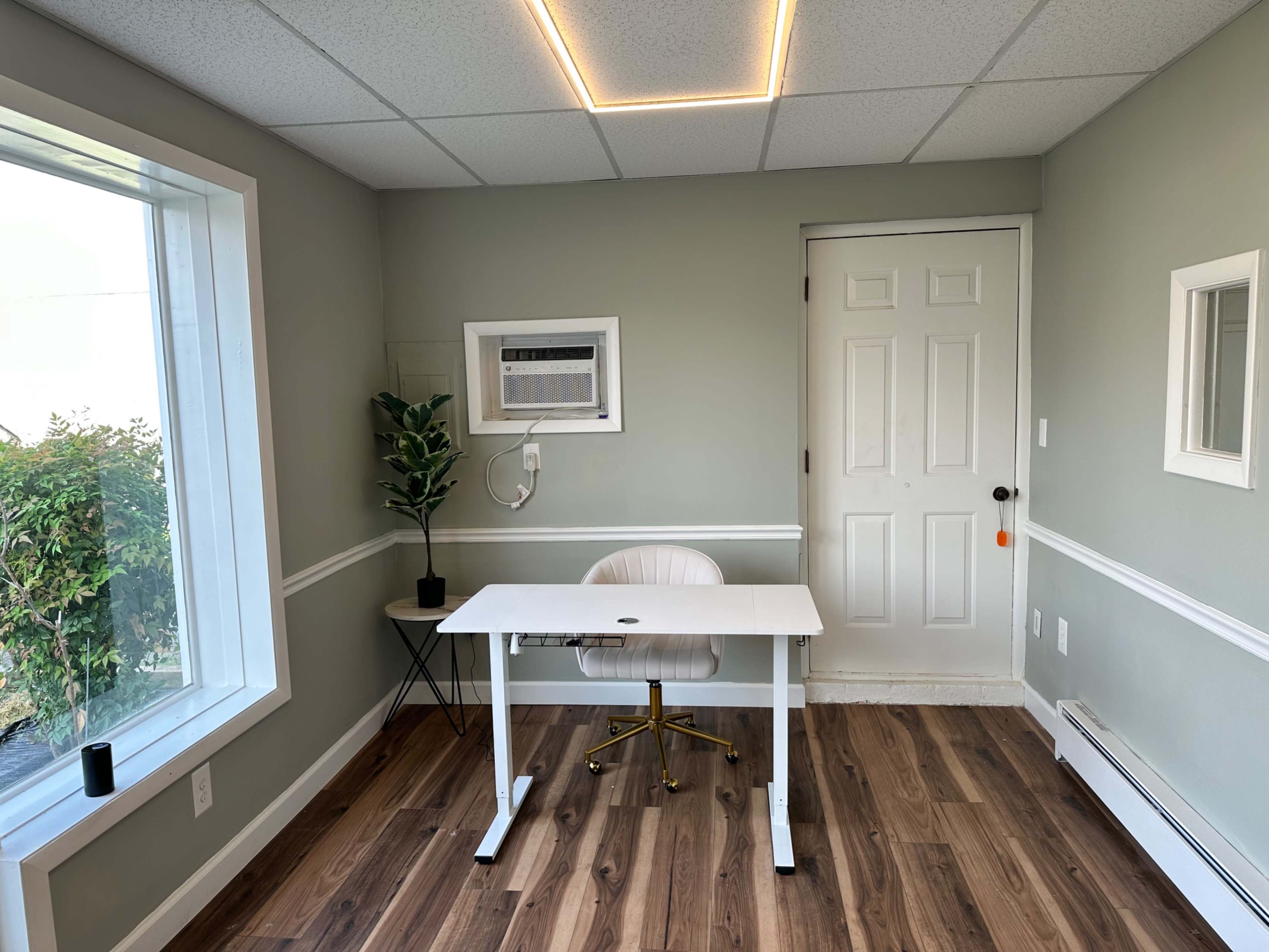 The image shows a small, minimalist office space featuring a white desk with a gold chair, an air conditioning unit, and green plants, along with two wall mirrors.