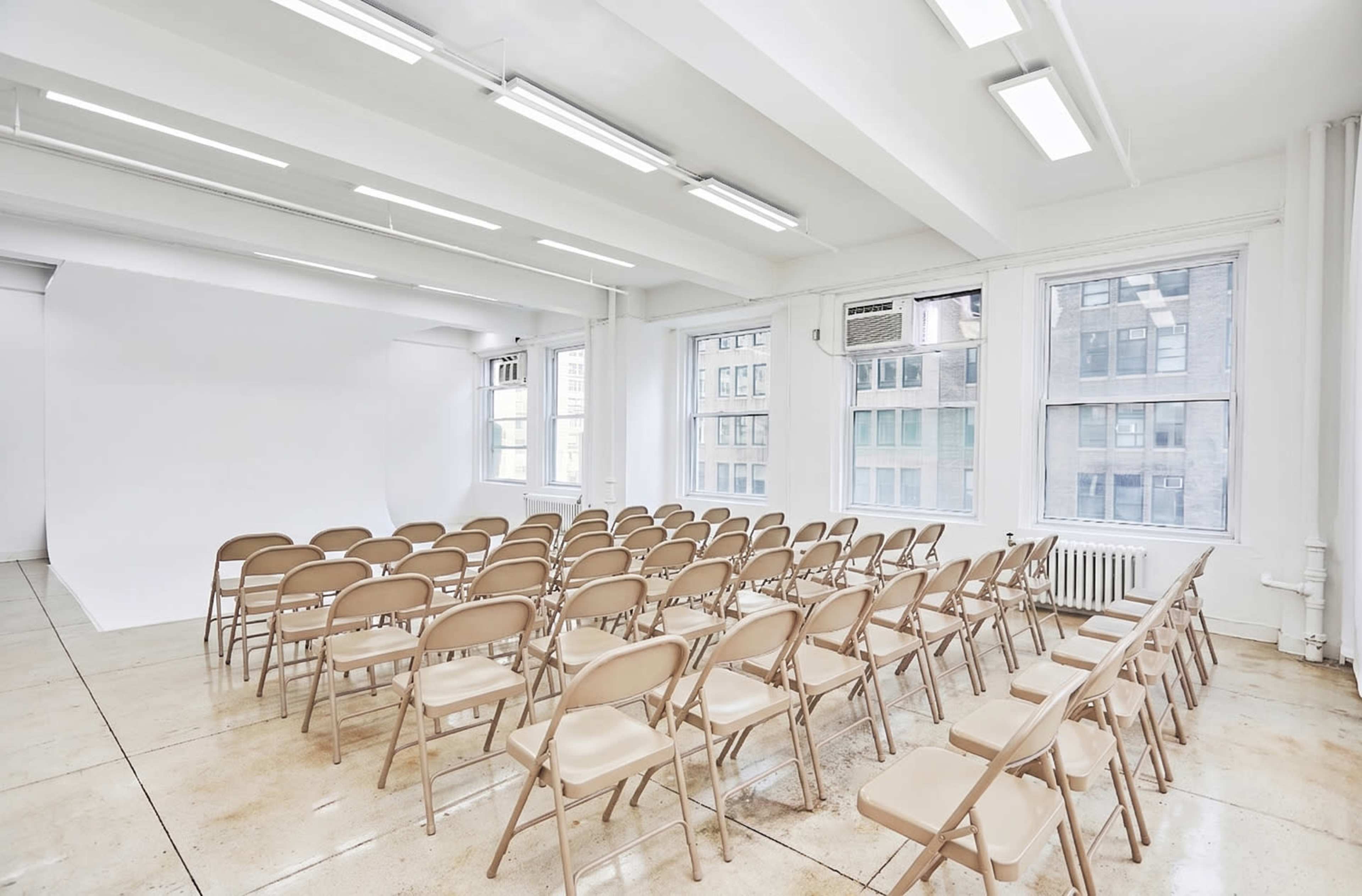 A bright, empty room features neatly arranged rows of beige folding chairs facing a blank white backdrop, with large windows allowing natural light to flow in.