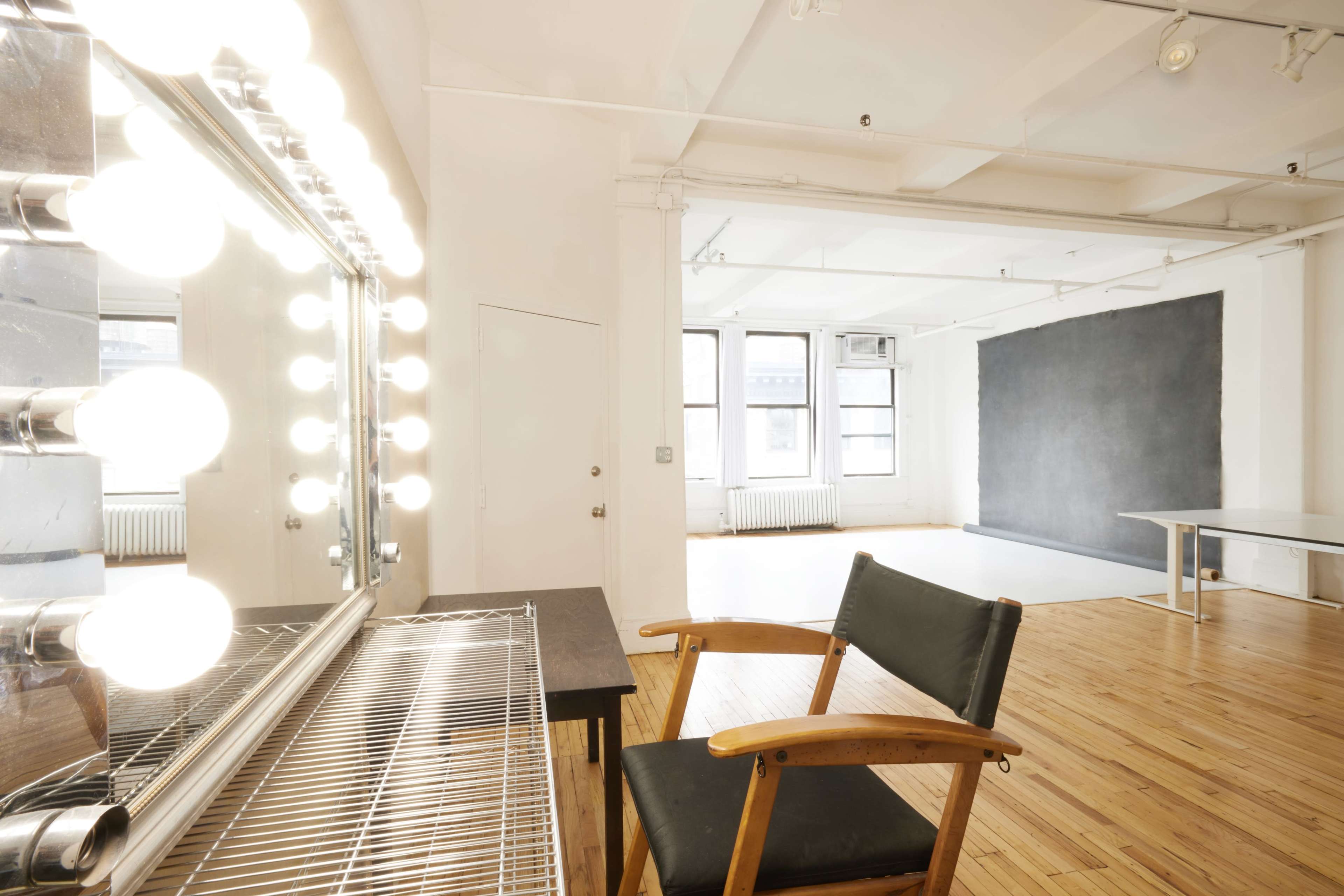 A bright, spacious room with a large mirror framed by lights, a wooden chair, and a black wall in the background.