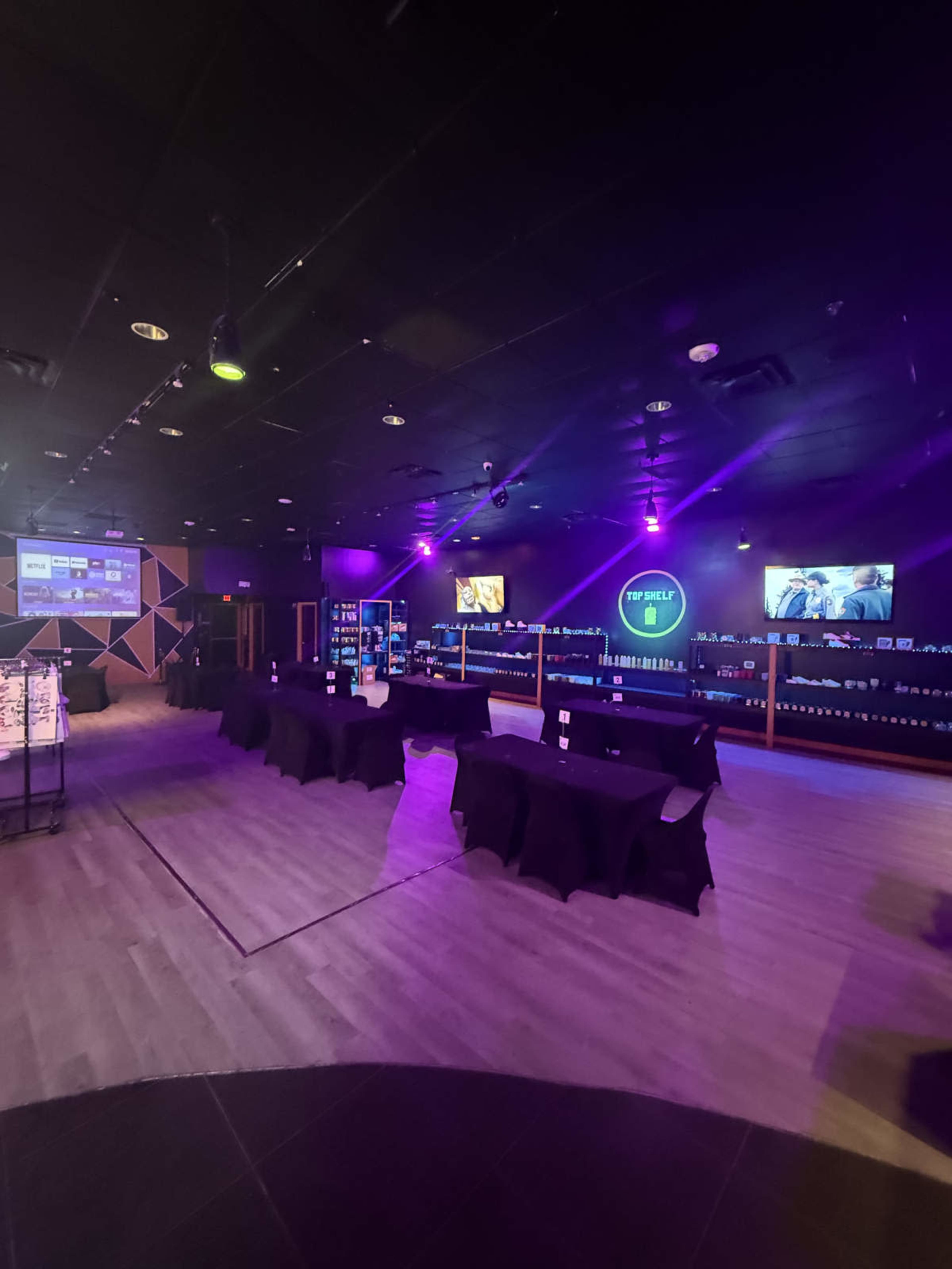 The image shows a dimly lit spacious room with several black tables set up, shelves lined with products in the background, and screens displaying content on the walls.