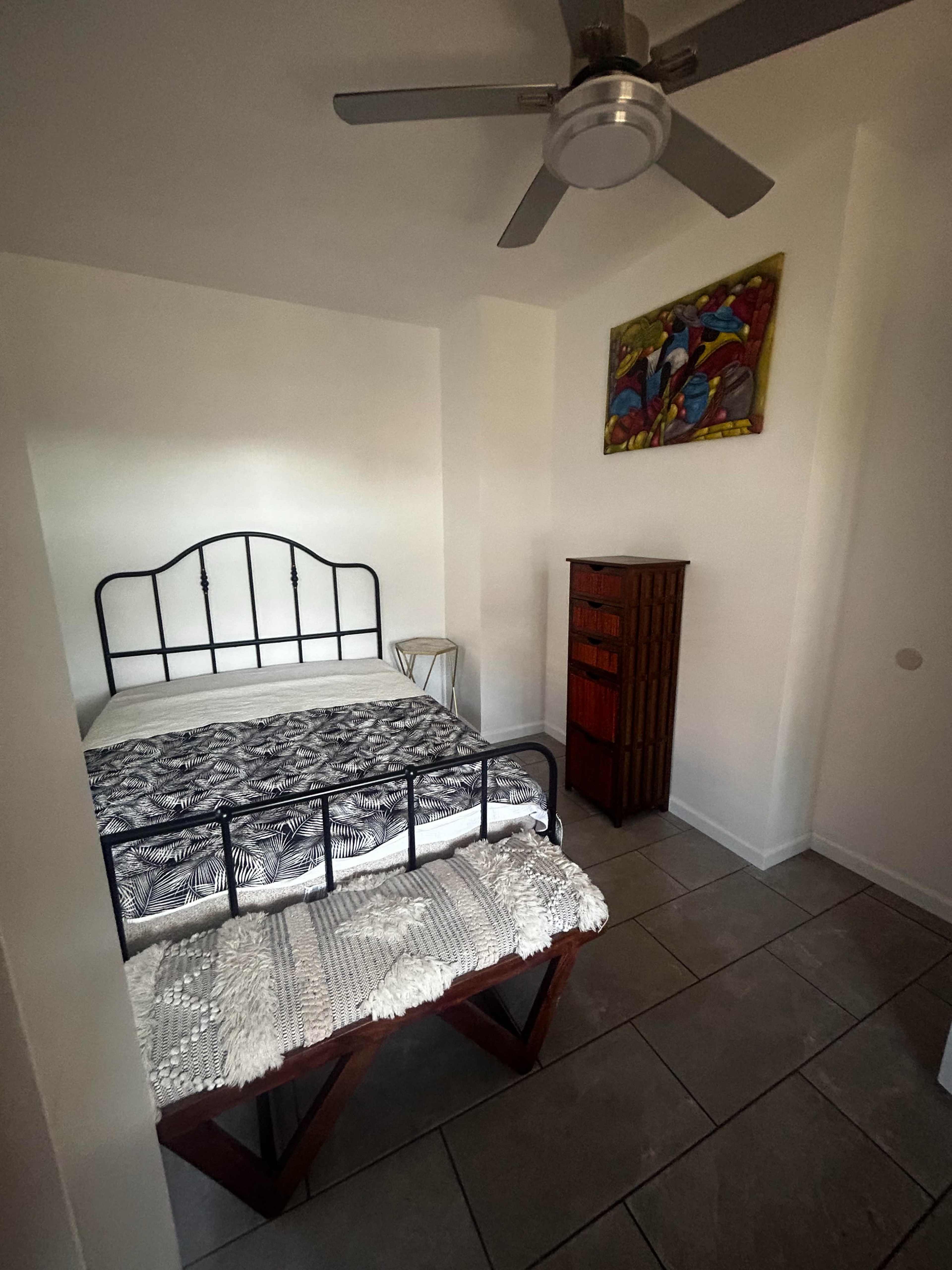 The image shows a small bedroom featuring a black metal bed, a white woven bench at the foot of the bed, a wooden dresser, and a ceiling fan, with a colorful framed artwork on the wall.