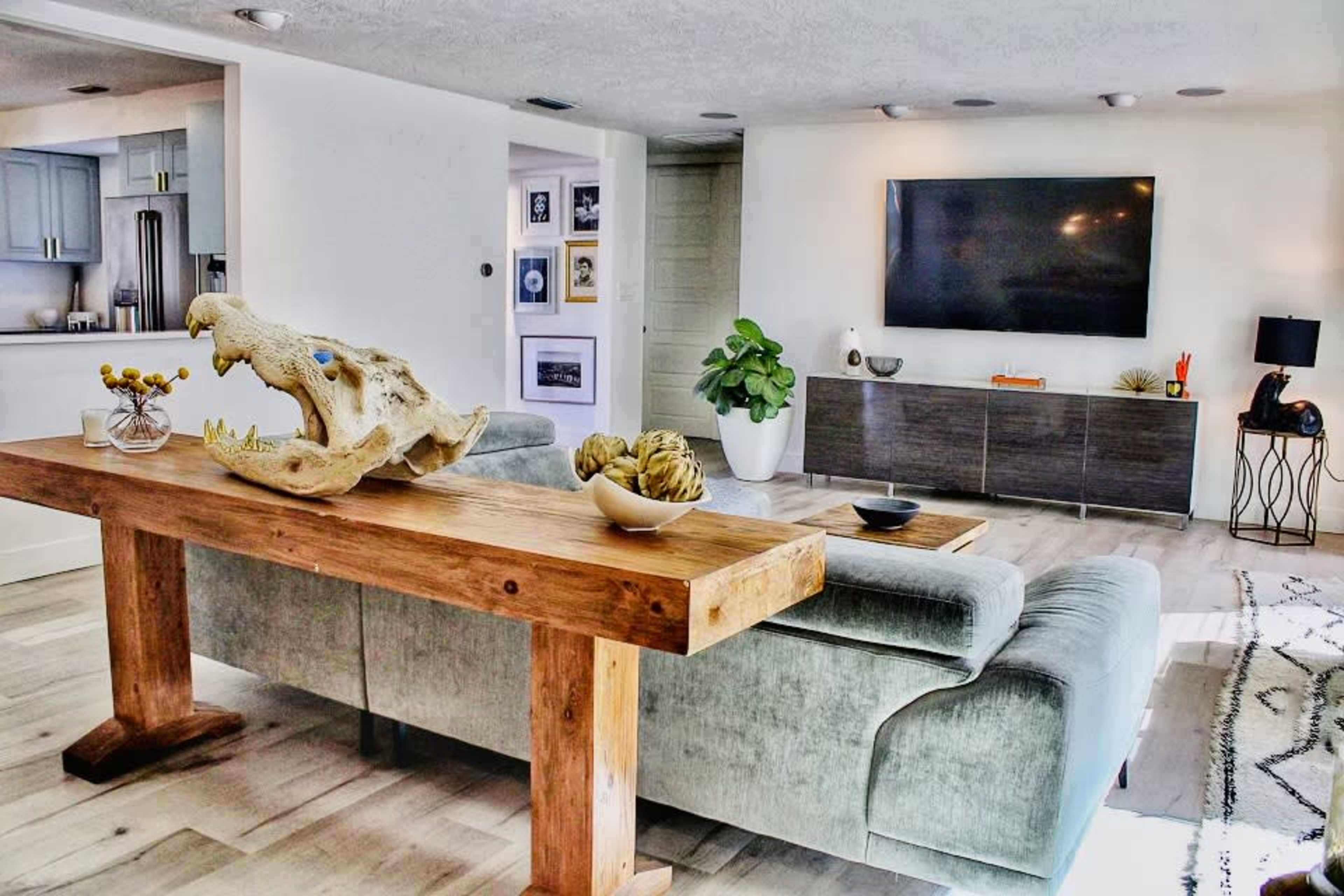 The image shows a modern living room with a gray sofa, a wooden dining table with decorative elements, a large TV mounted on the wall, and a potted plant in the corner.