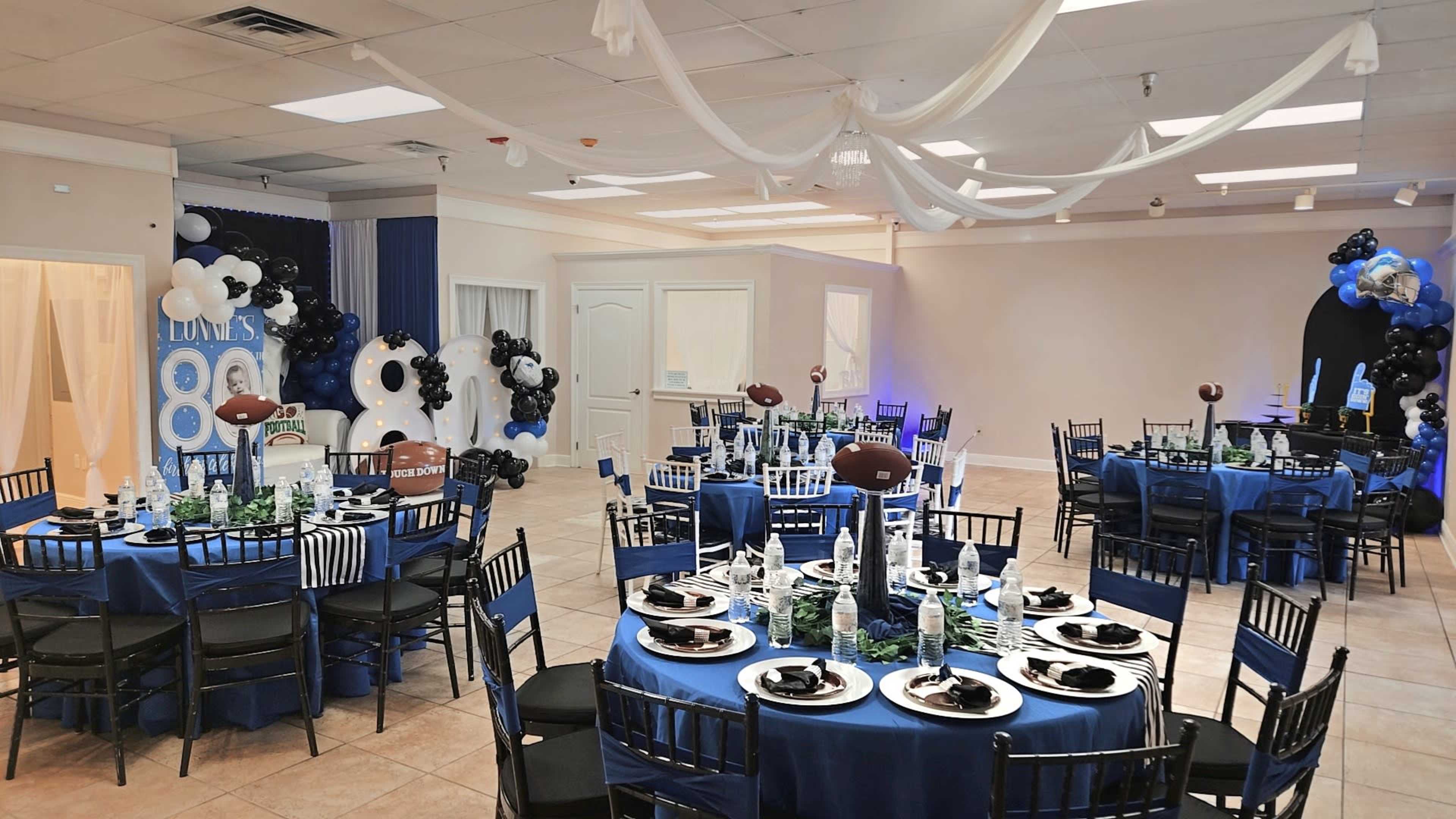 The image shows a decorated indoor event space with tables set in blue and black, featuring football-themed centerpieces and party decorations.