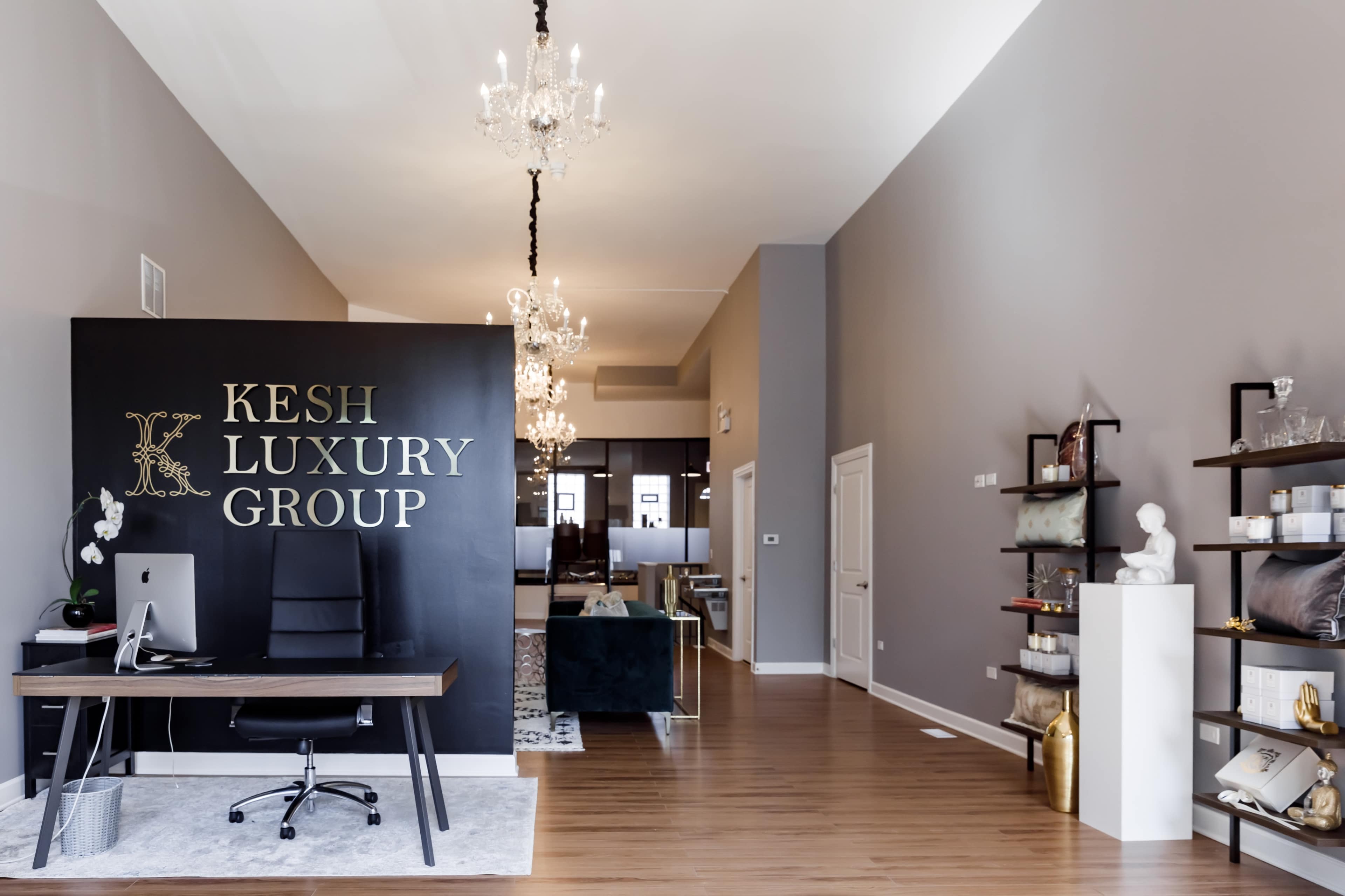 A modern office space featuring a black reception desk with the name "KESH LUXURY GROUP," elegant chandeliers hanging from the ceiling, and a spacious area with furniture arranged for a professional setting.