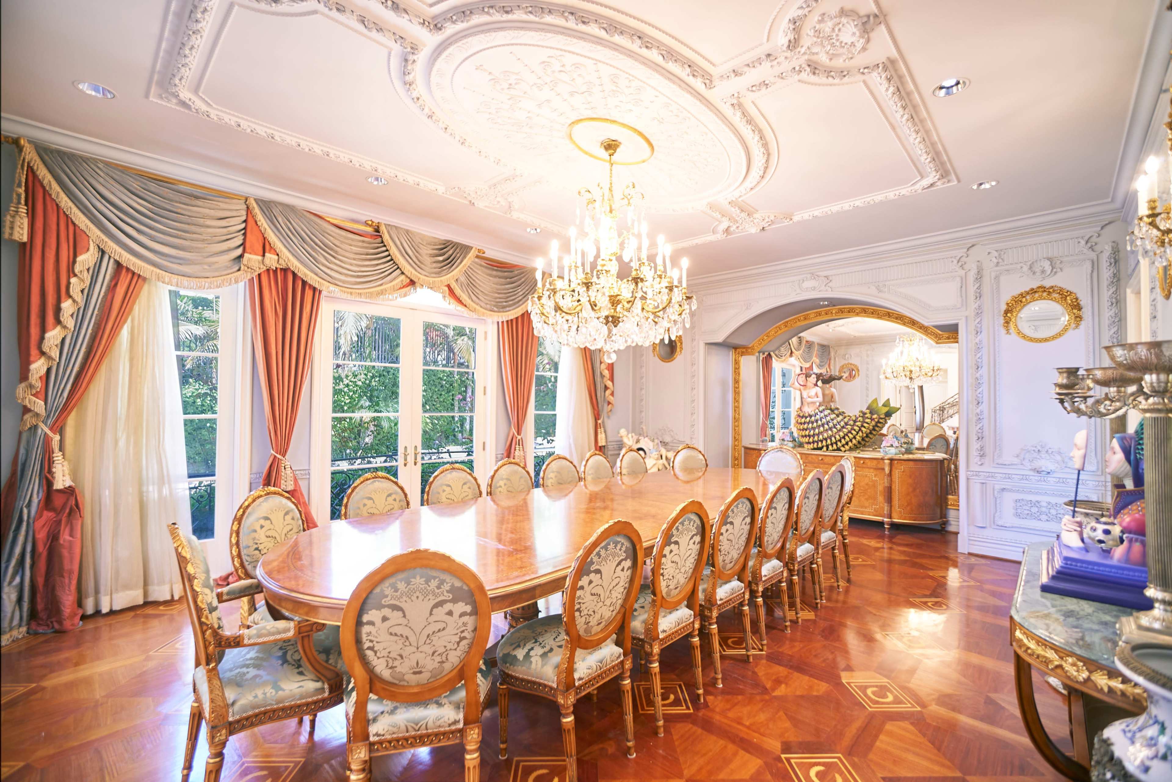 A large dining room features an ornate chandelier, a long wooden table surrounded by upholstered chairs, and elaborate drapery framing large windows.