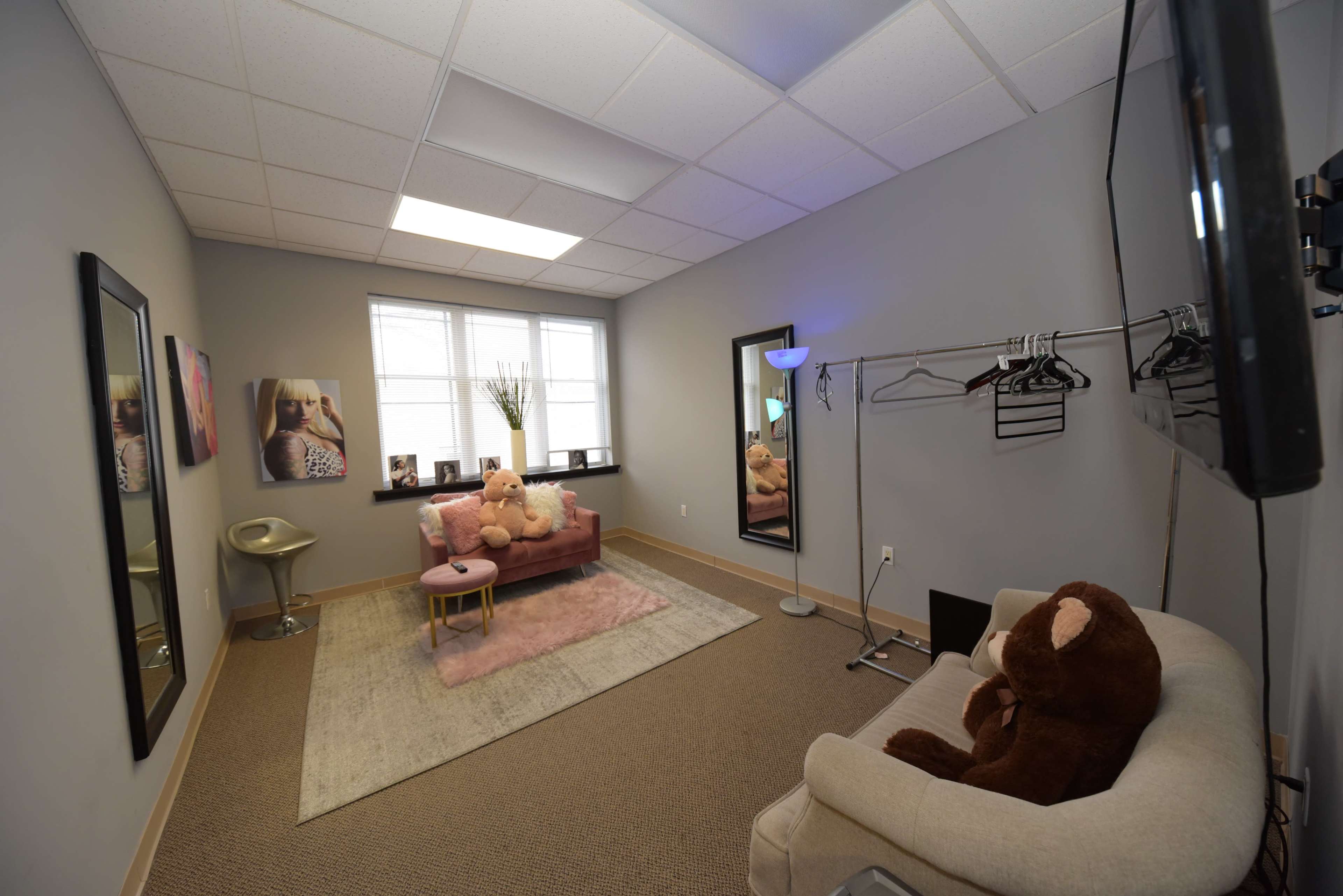A small, well-lit room featuring a pink sofa, a white chair, a small table, and two mirrors, with a clothing rack and a television on the wall.