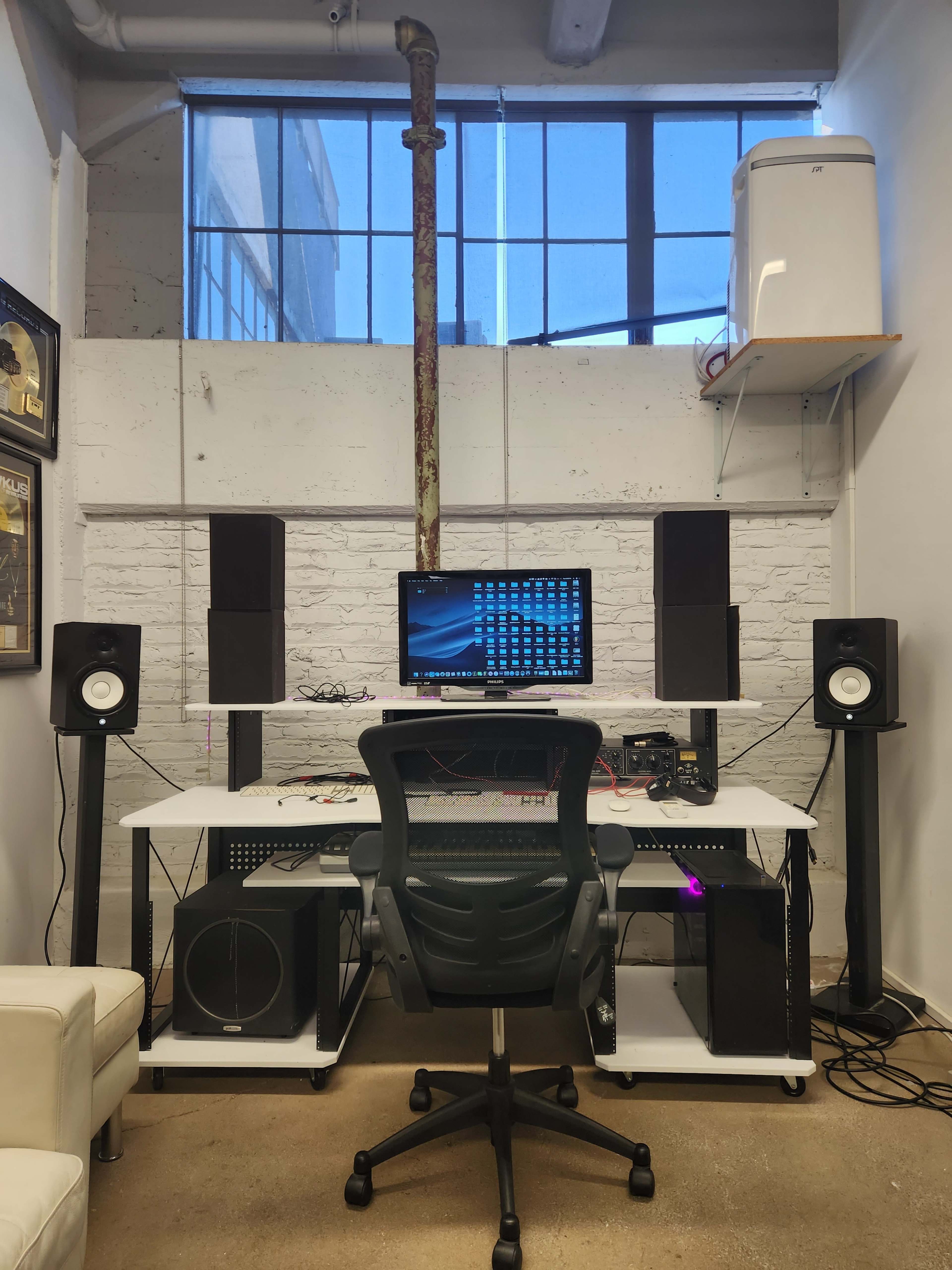 A computer desk with a monitor, speakers, and audio equipment is positioned against a wall with a window and a visible pipe in the background.