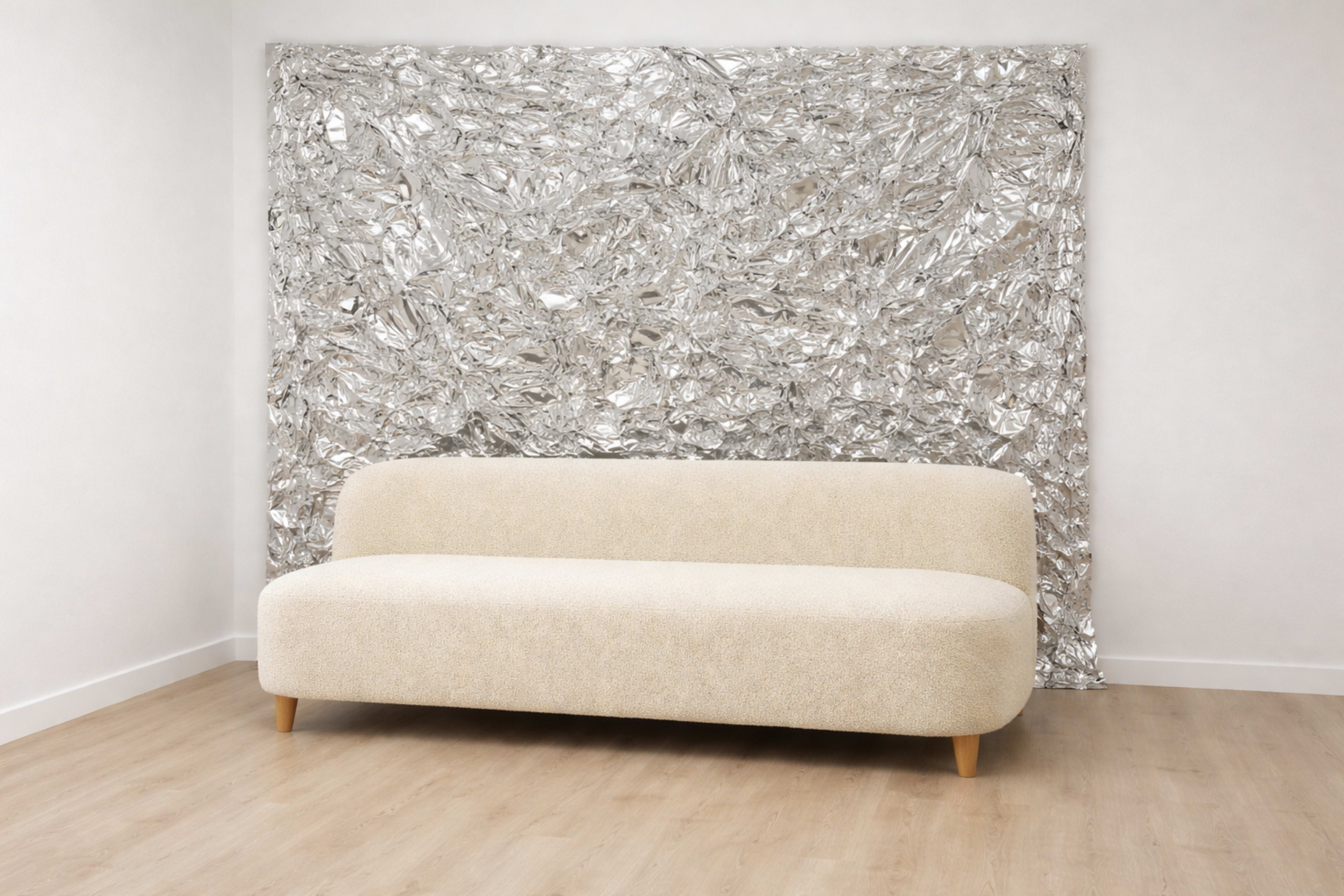 A beige, curved sofa is positioned in front of a shiny, crumpled metallic wall art in a room with light wooden flooring.