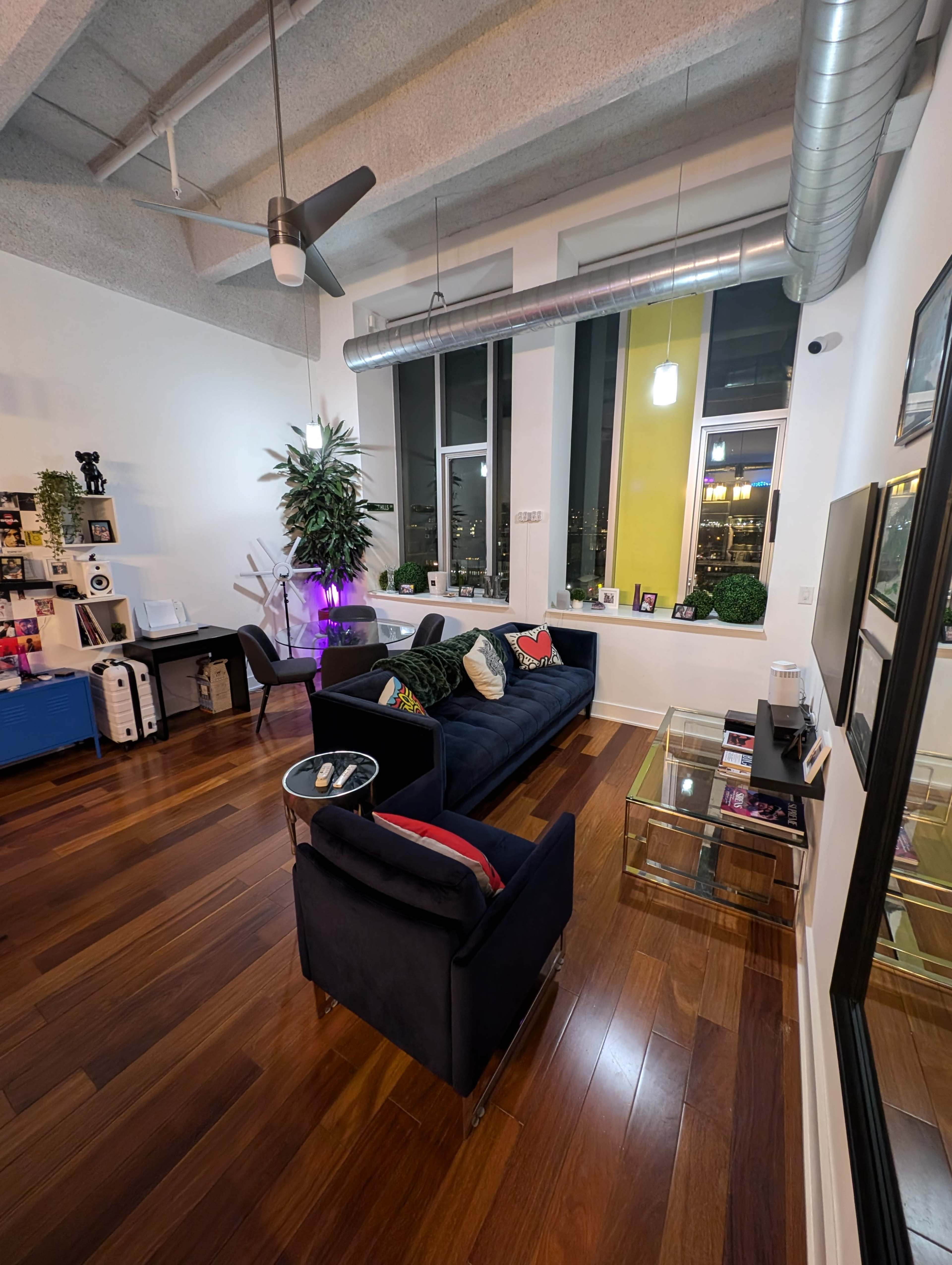 The image shows a modern living room with a navy blue sofa, two chairs, a coffee table, and large windows featuring indoor plants and various decorative items.
