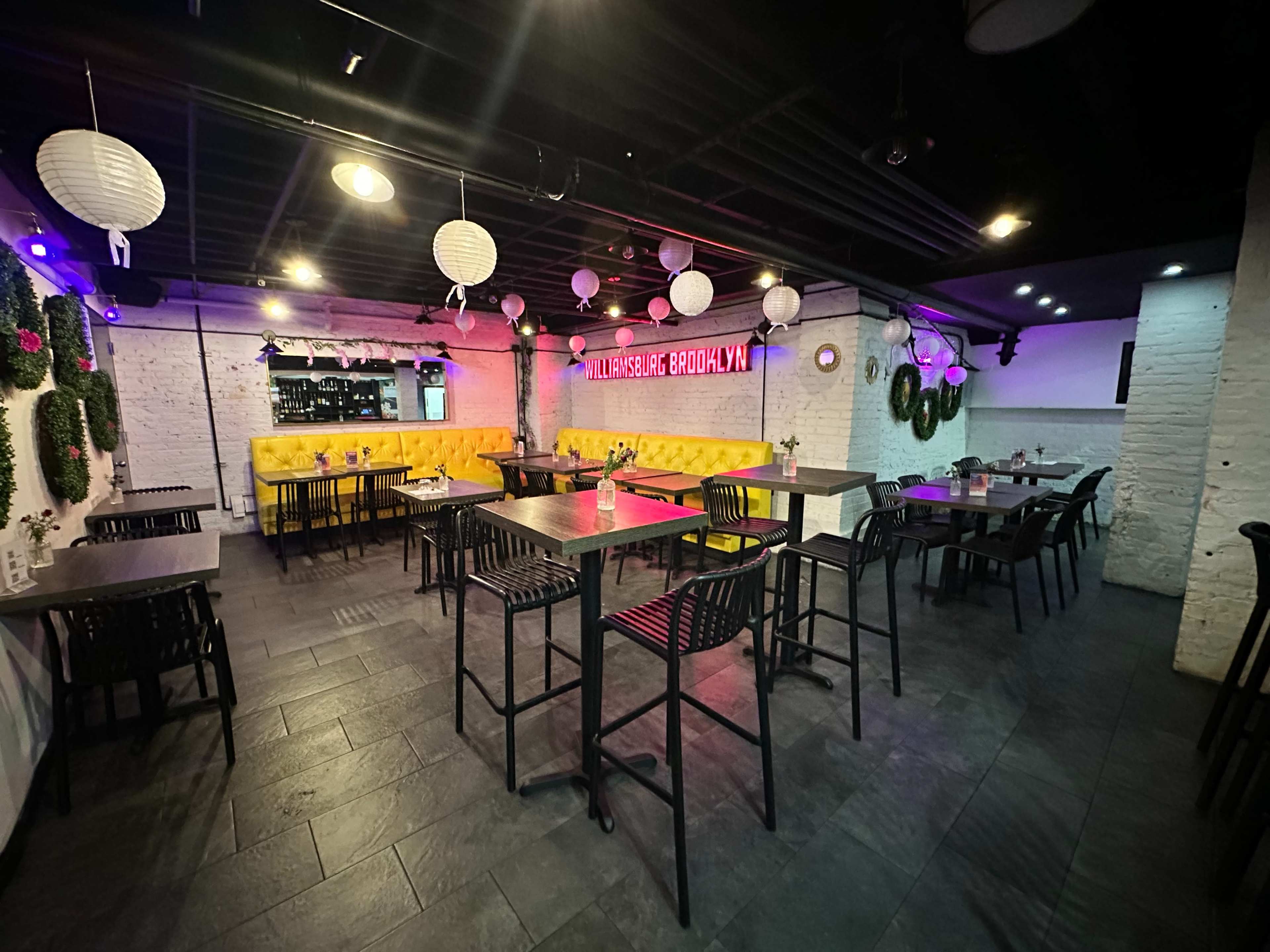 A dimly lit restaurant interior with black tables and chairs, colorful decor, and a bright neon sign on the wall.