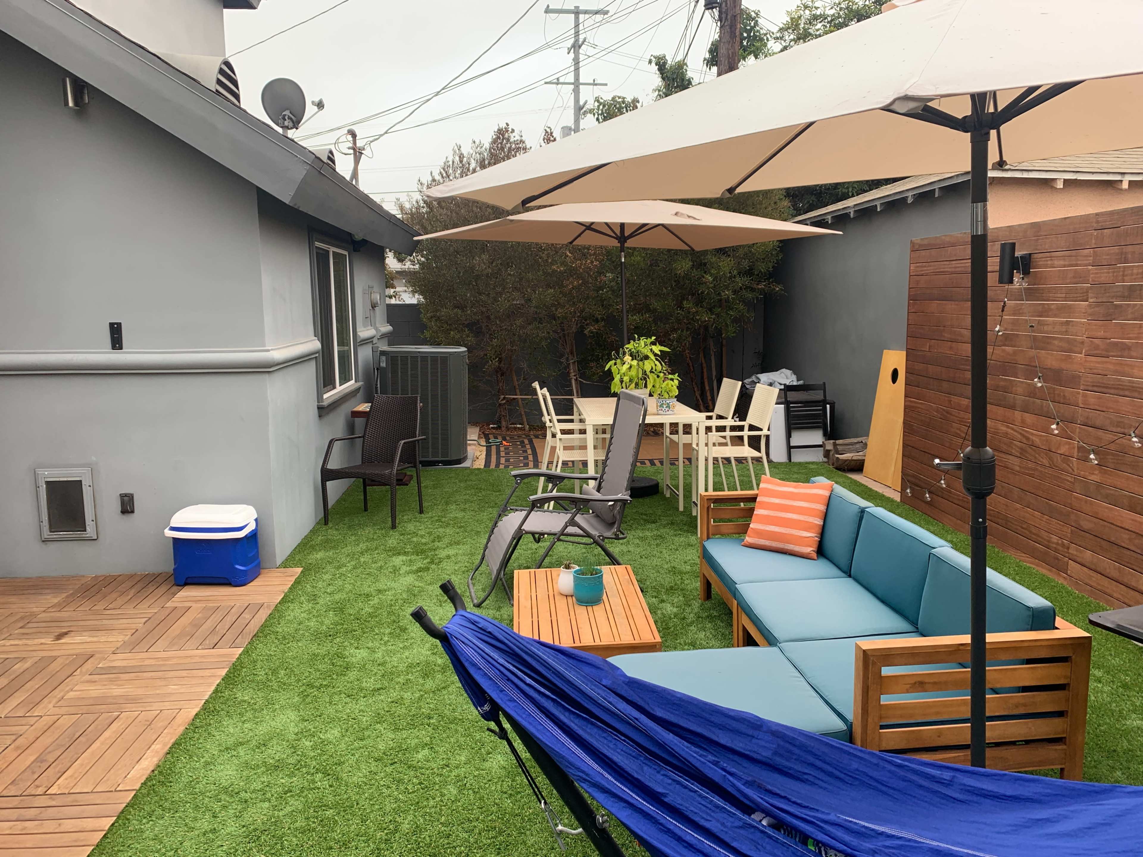 A backyard features a patio with a seating area, dining table, and umbrellas, surrounded by artificial grass and wooden walls.