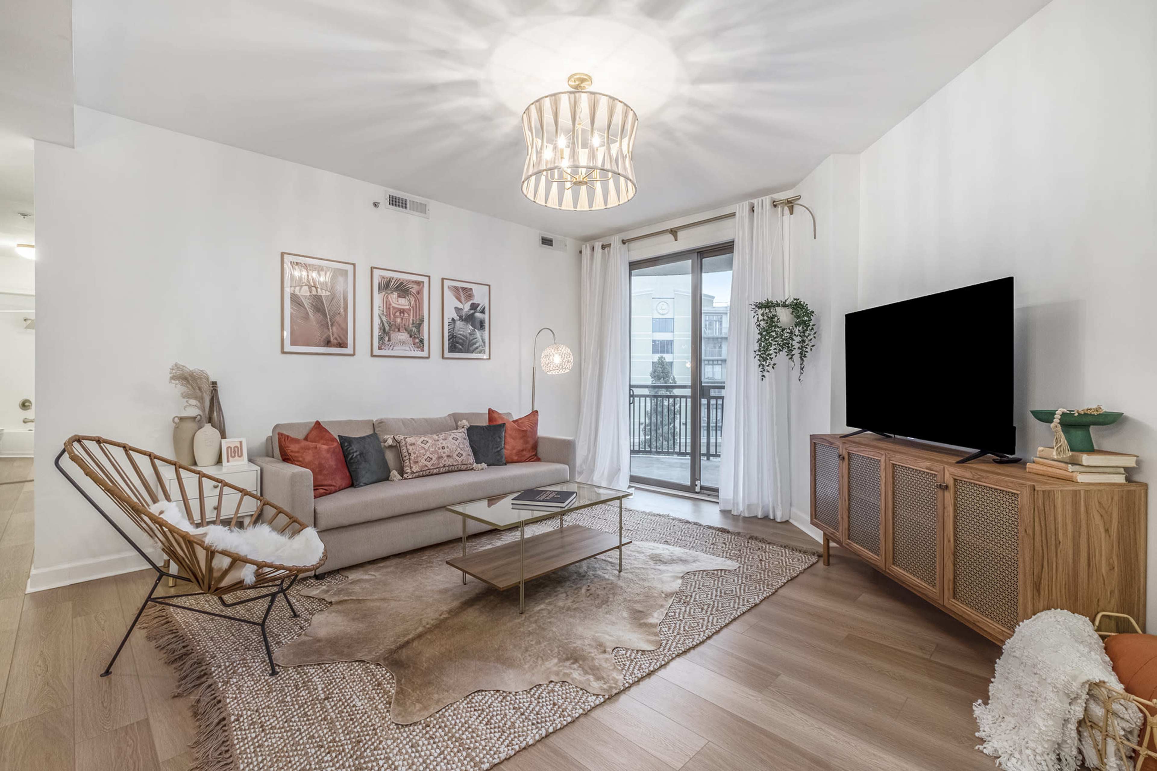 A modern living room features a sofa with decorative pillows, a coffee table, a large TV, a light fixture, and sliding glass doors opening to a balcony.