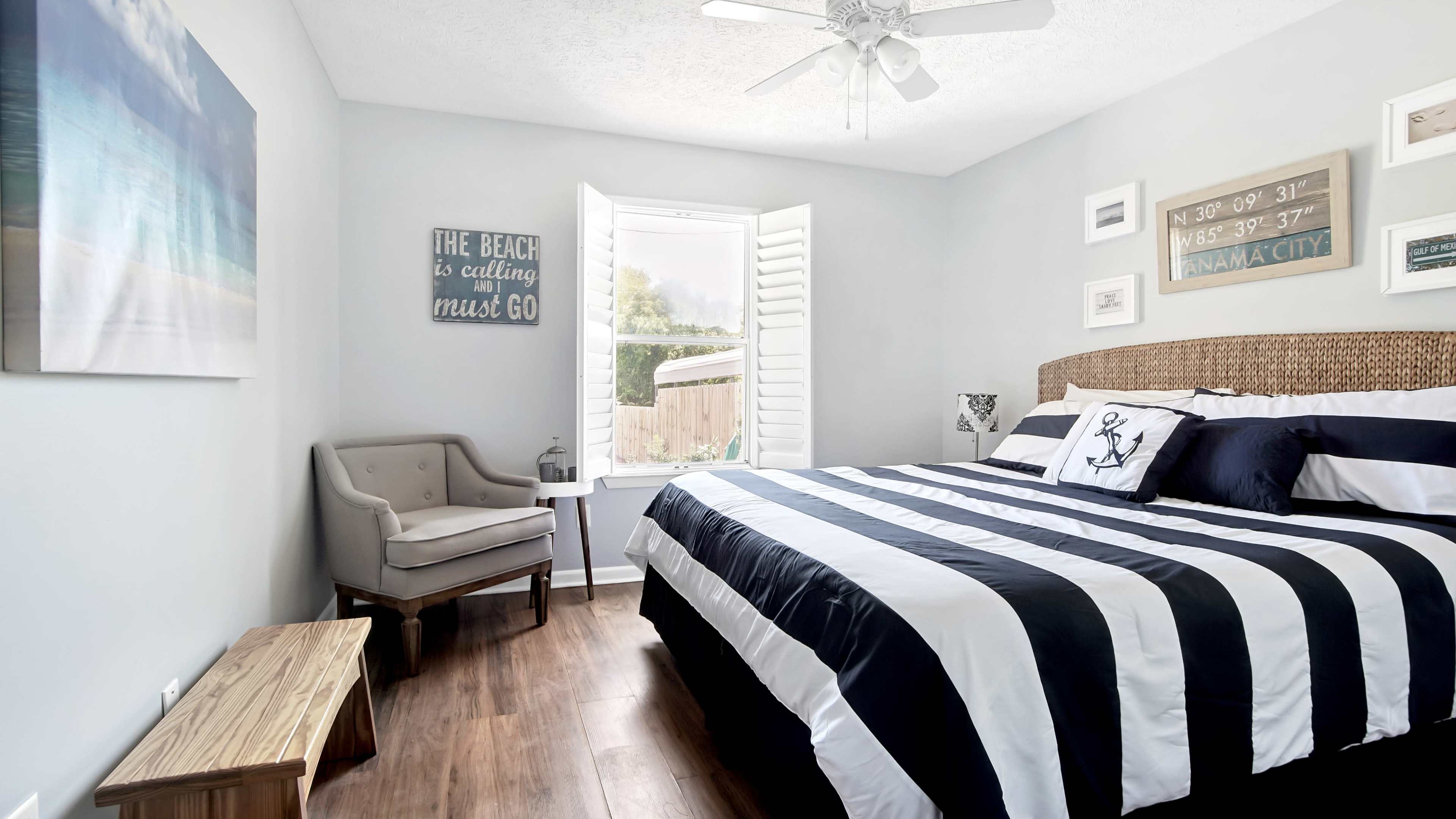 A bedroom features a striped bedspread, a chair beside a window with shutters, and beach-themed decor on the walls.