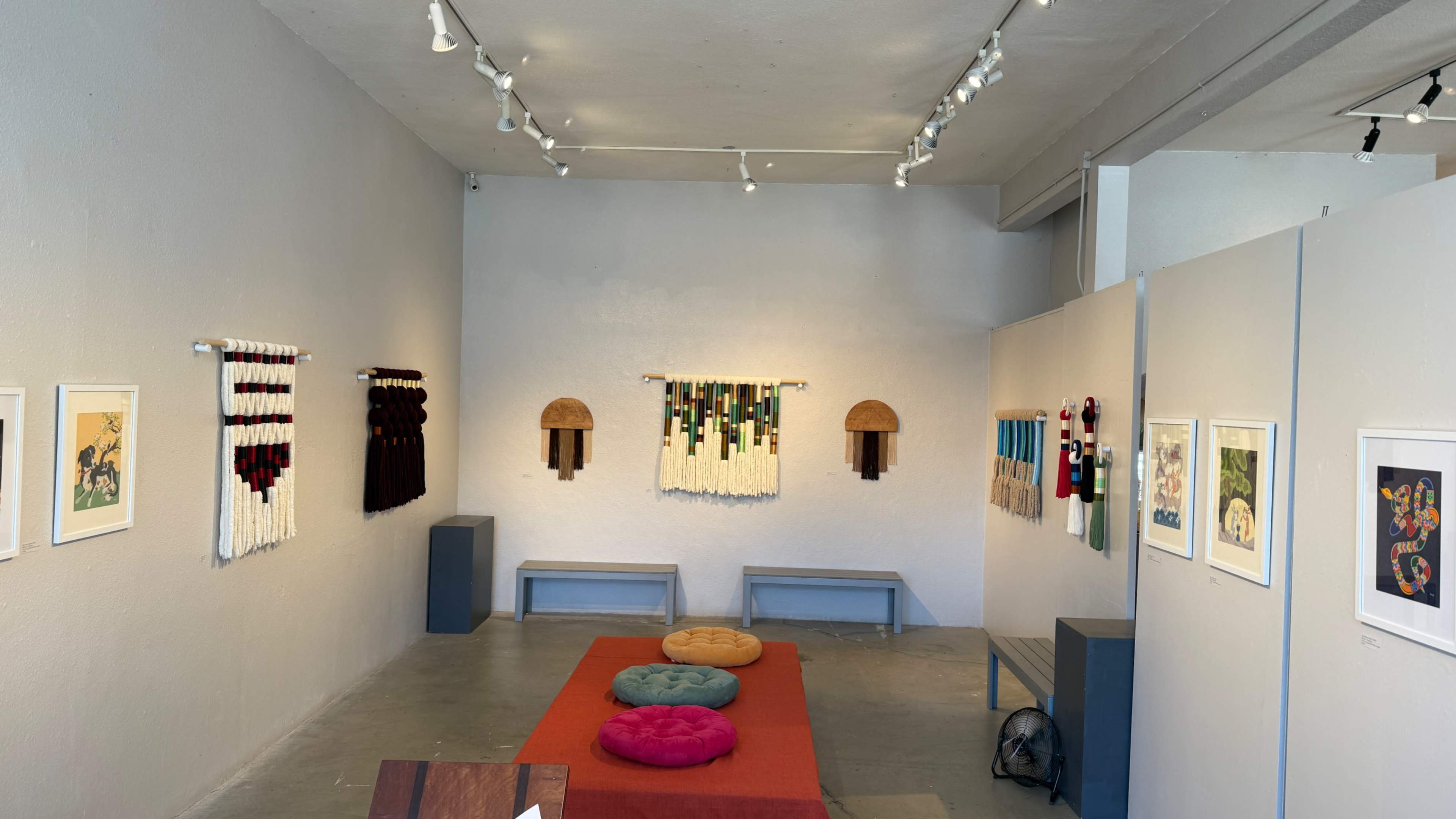 The image shows an art gallery with colorful textile wall hangings and various artworks displayed on the walls, along with seating cushions on a red carpet.