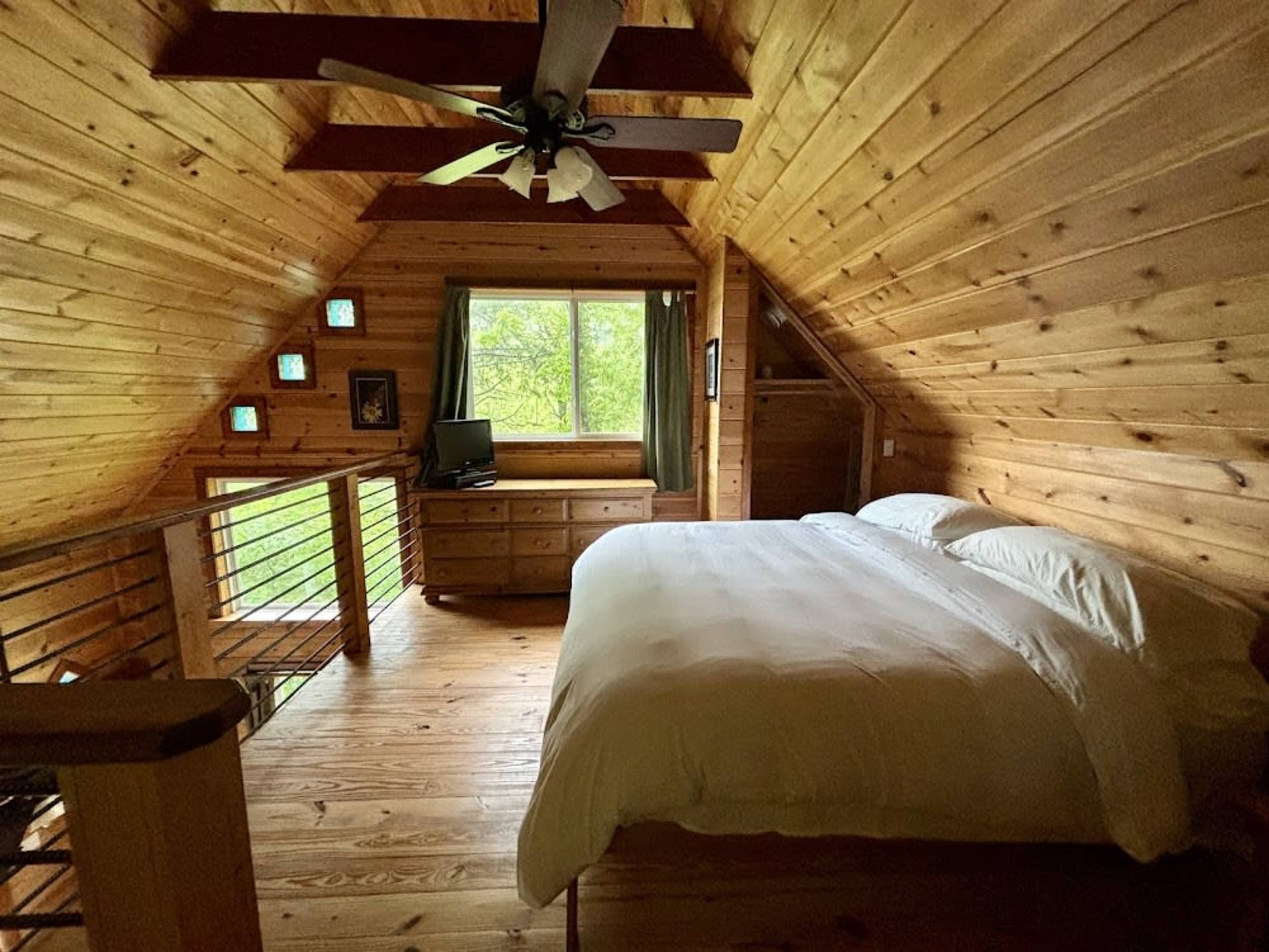 The image shows a wooden loft bedroom featuring a bed, dresser, and natural light coming through a large window.