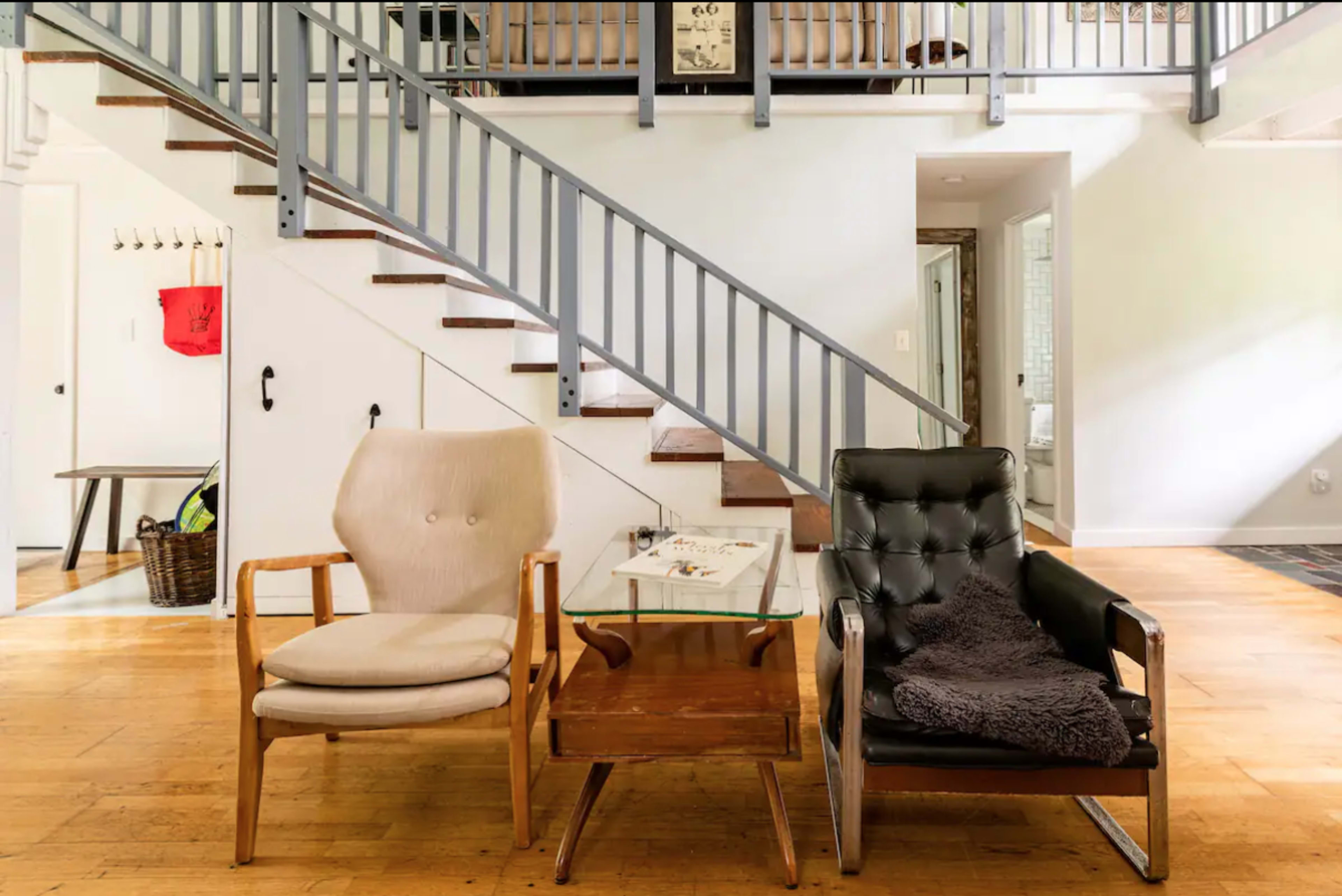 Two armchairs flank a glass coffee table in a well-lit, open living space with a staircase in the background.