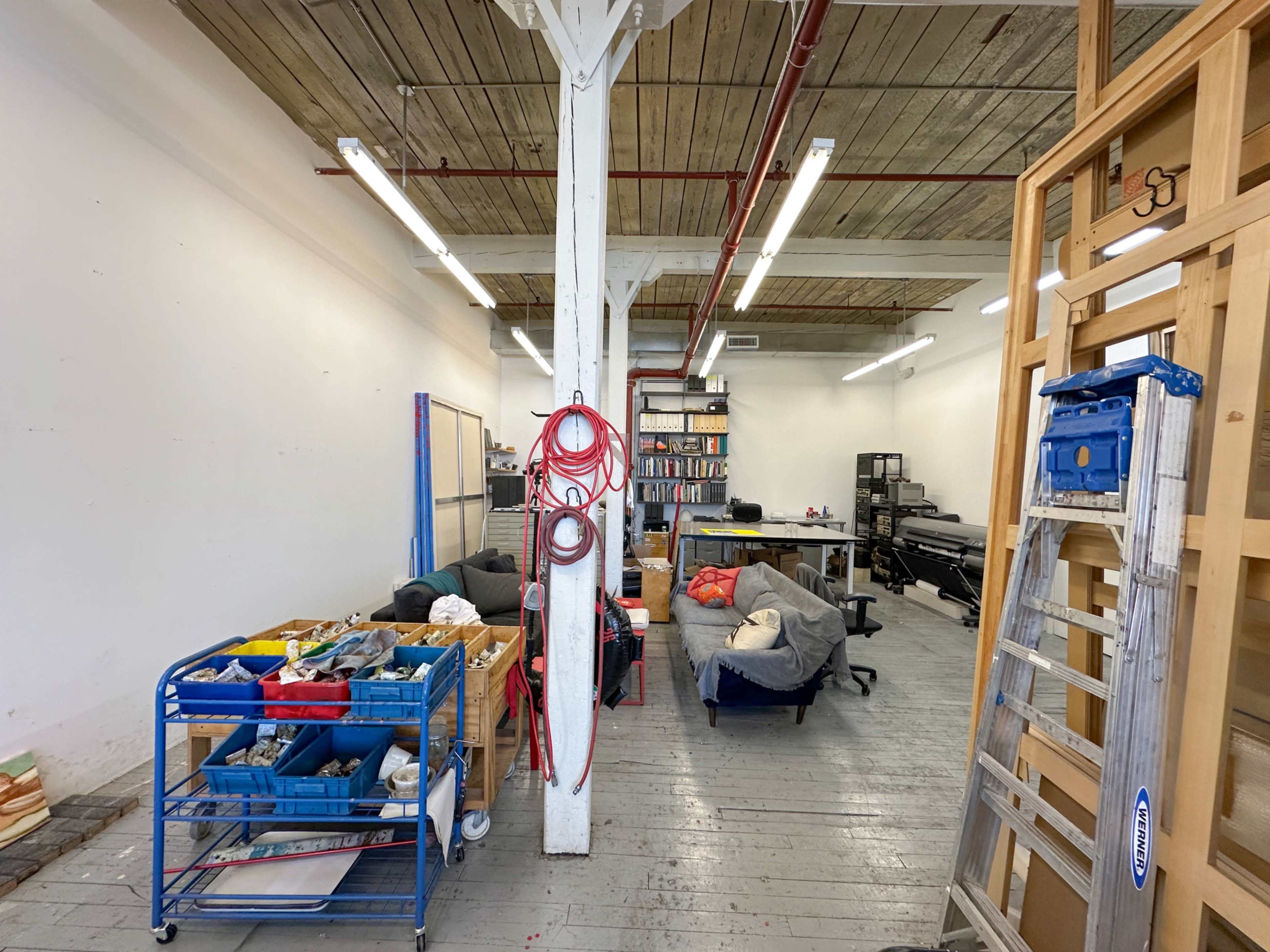 The image shows a spacious workshop or studio with wooden beams, various tools and materials organized on carts, and seating areas with bean bags.