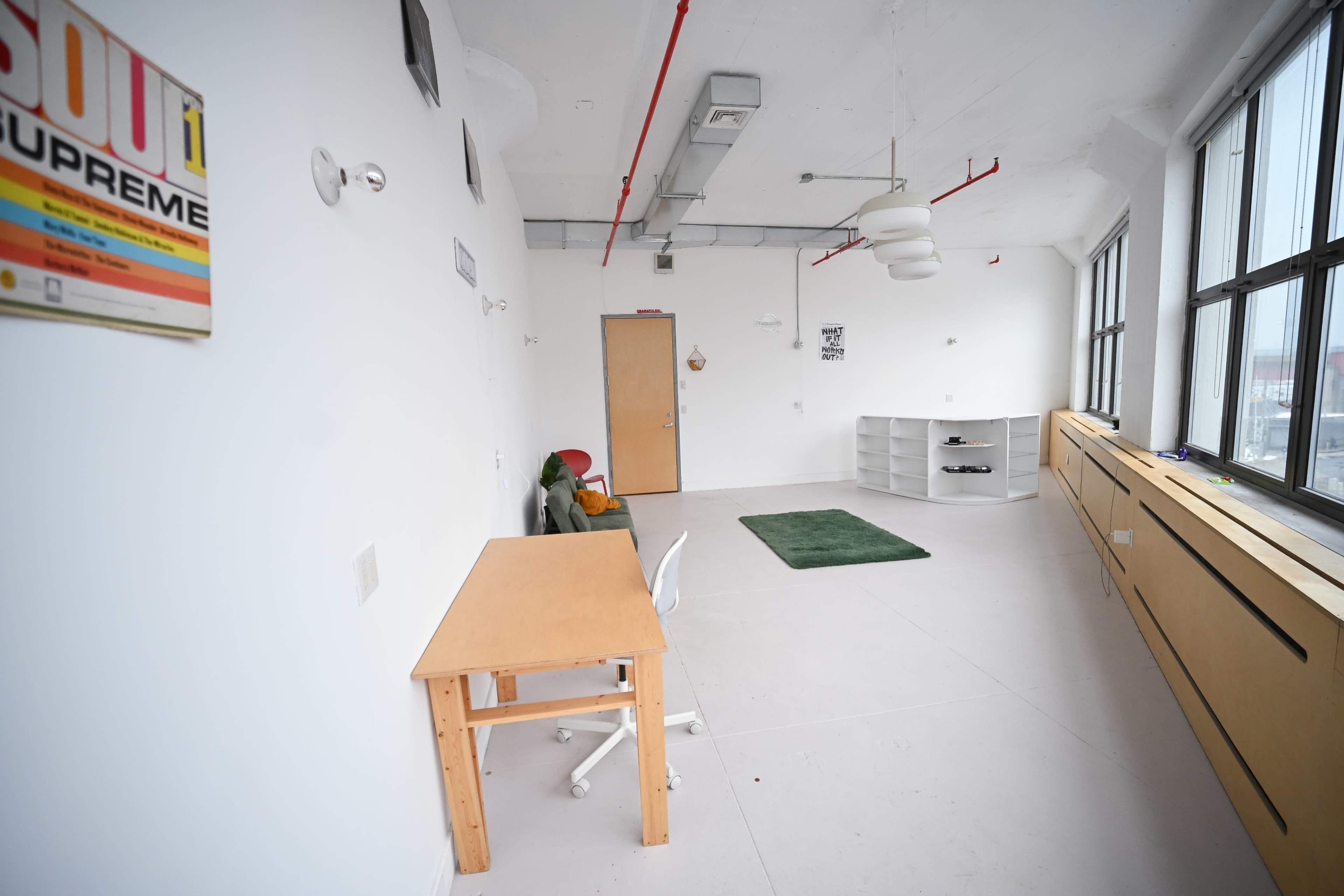 The image shows a minimalist, empty room with white walls, a wooden desk and chair, a green rug on the floor, and a shelf unit against one wall.