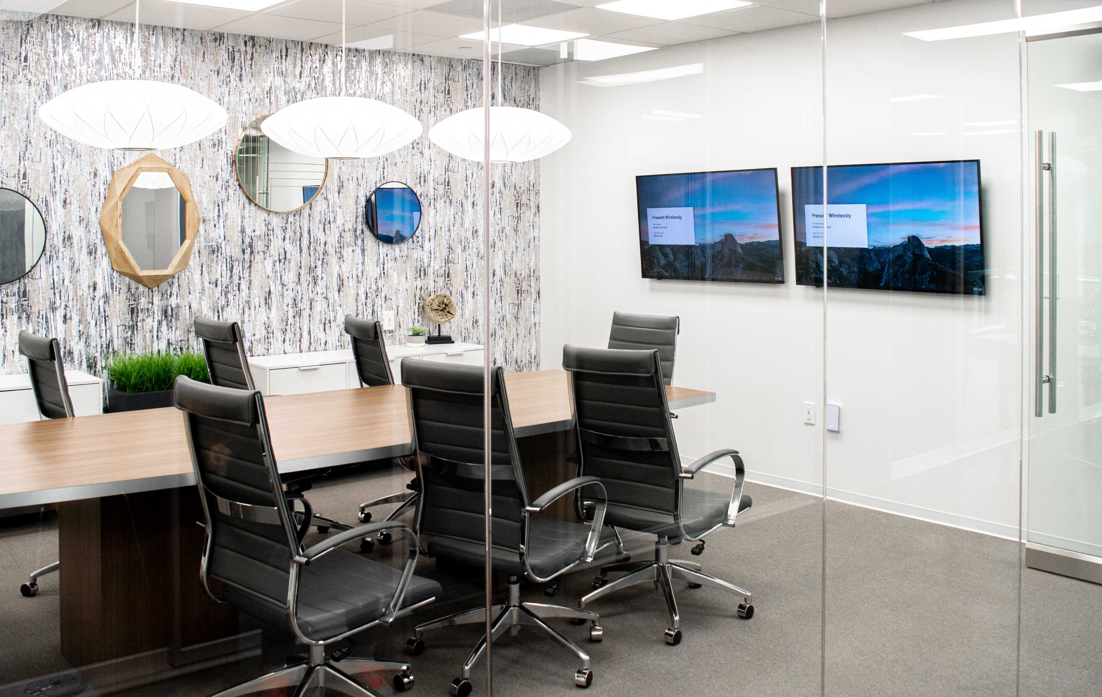 The image shows a modern conference room with a long wooden table, black leather chairs, mirrored wall decor, and two screens displaying presentations.