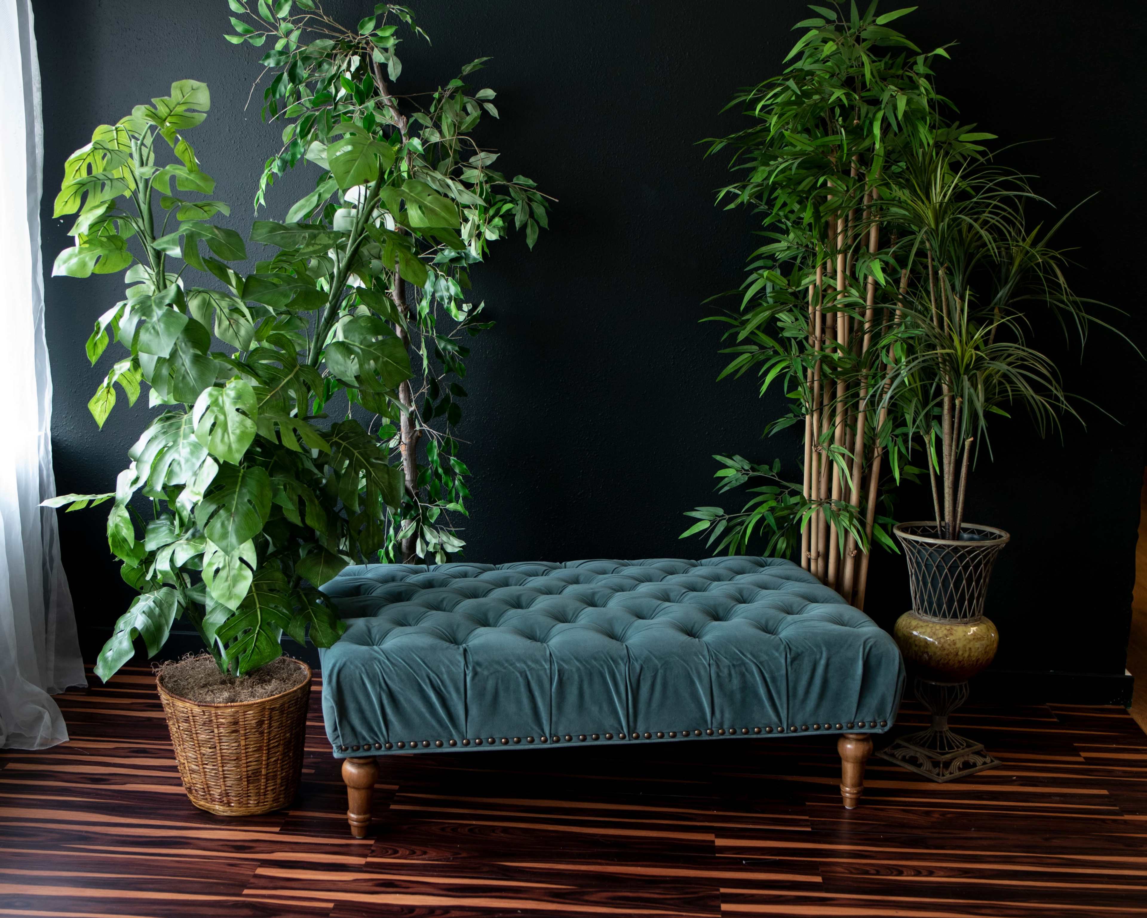 A tufted teal ottoman is positioned between two potted plants against a black wall.