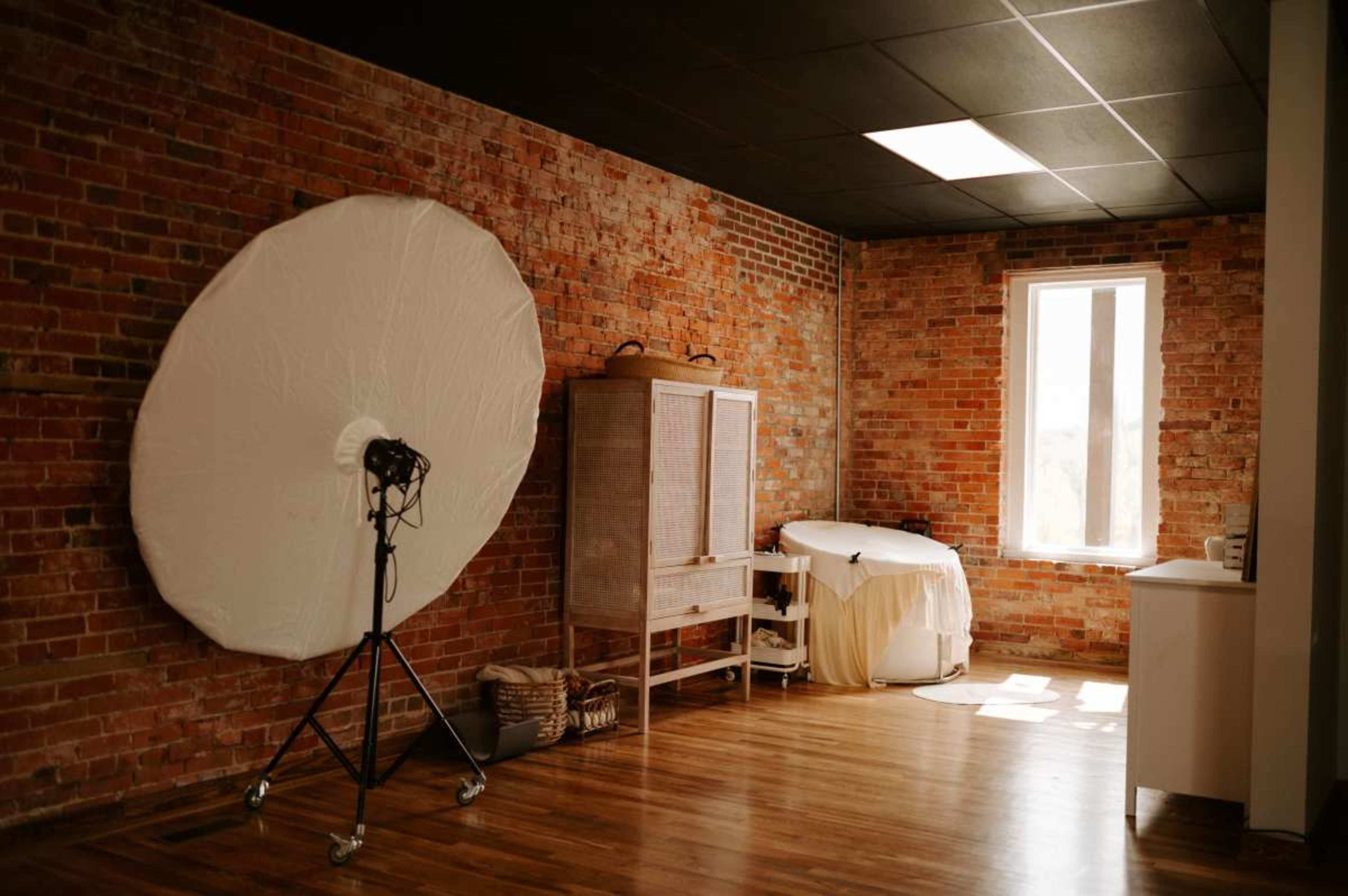 A spacious interior with exposed brick walls, featuring a large reflector on a stand, a wooden cabinet, a bathtub, and a window allowing natural light to enter.