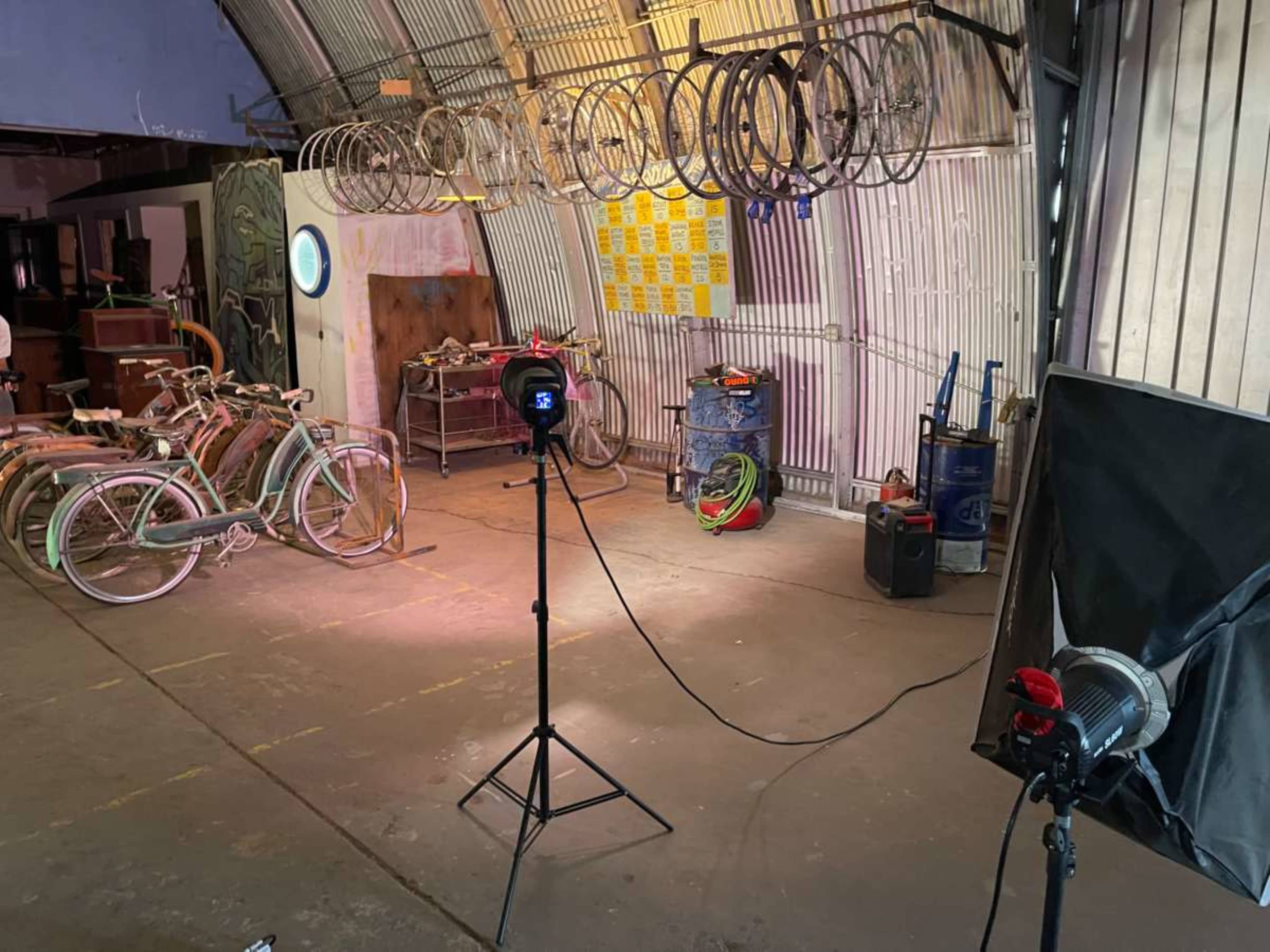 The image shows an industrial workshop with bicycles lined against the walls, multiple bicycle rims hanging from the ceiling, and studio lights set up for photography.