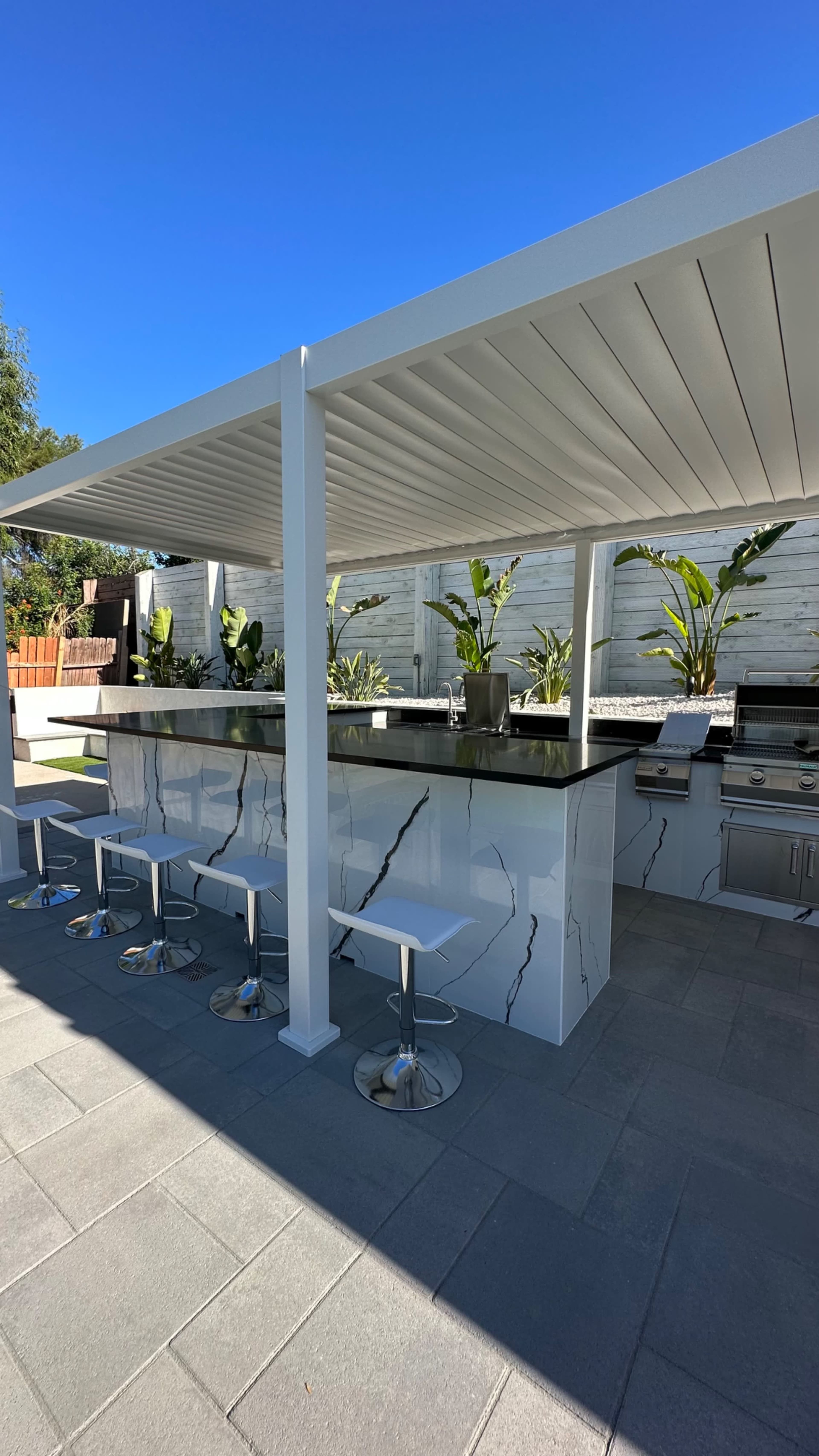 An outdoor kitchen area with a large countertop, bar stools, and built-in appliances beneath a covered structure.