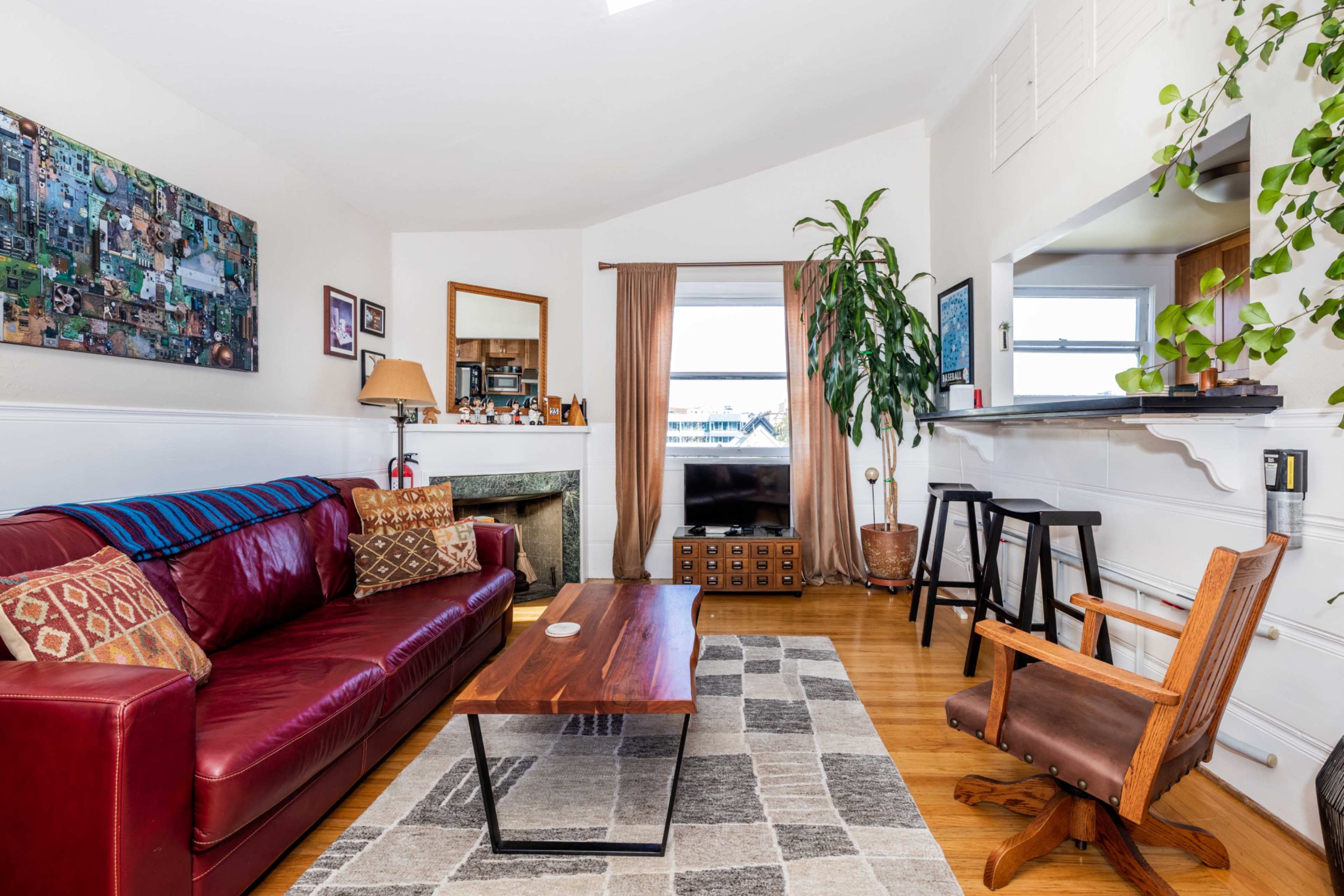 The image shows a cozy living room featuring a red leather sofa, a wooden coffee table, a large window with curtains, and various plants, with a television visible in the background.