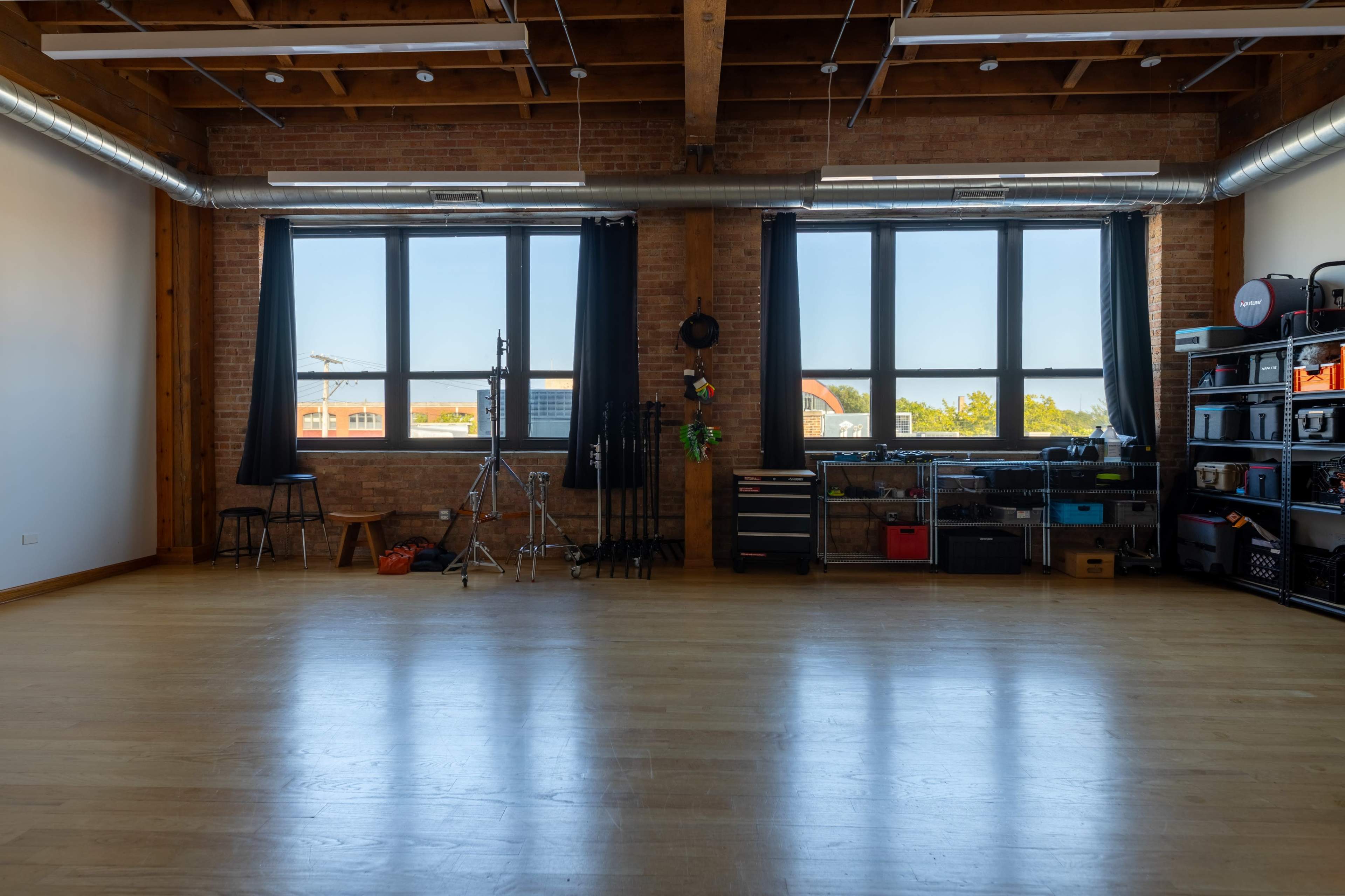 An empty studio space with large windows, wooden beams, and various photography equipment organized along one wall.