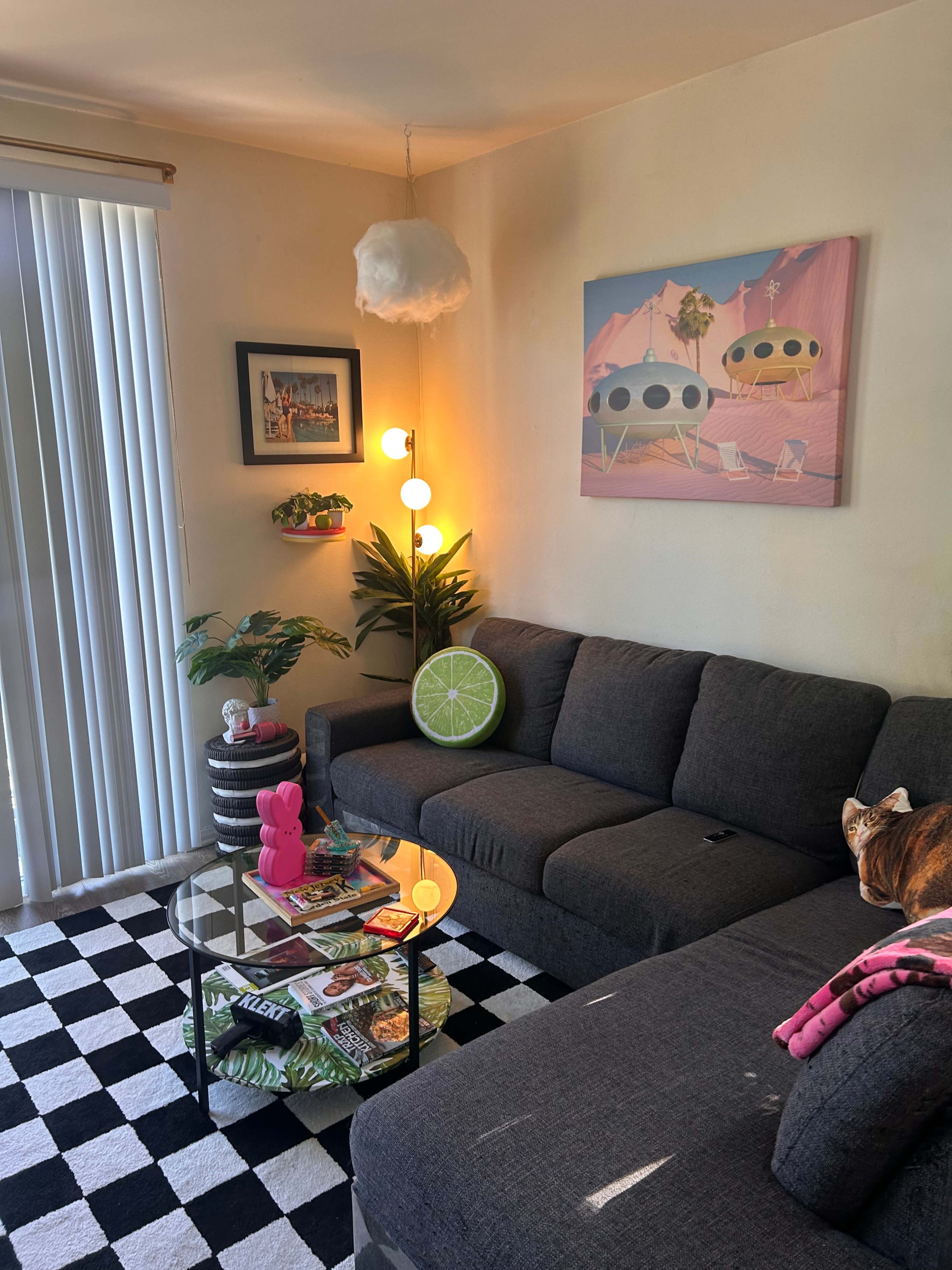 A modern living room features a dark gray sectional sofa, a round coffee table with magazines, a colorful wall art piece, and vertical blinds allowing natural light in.
