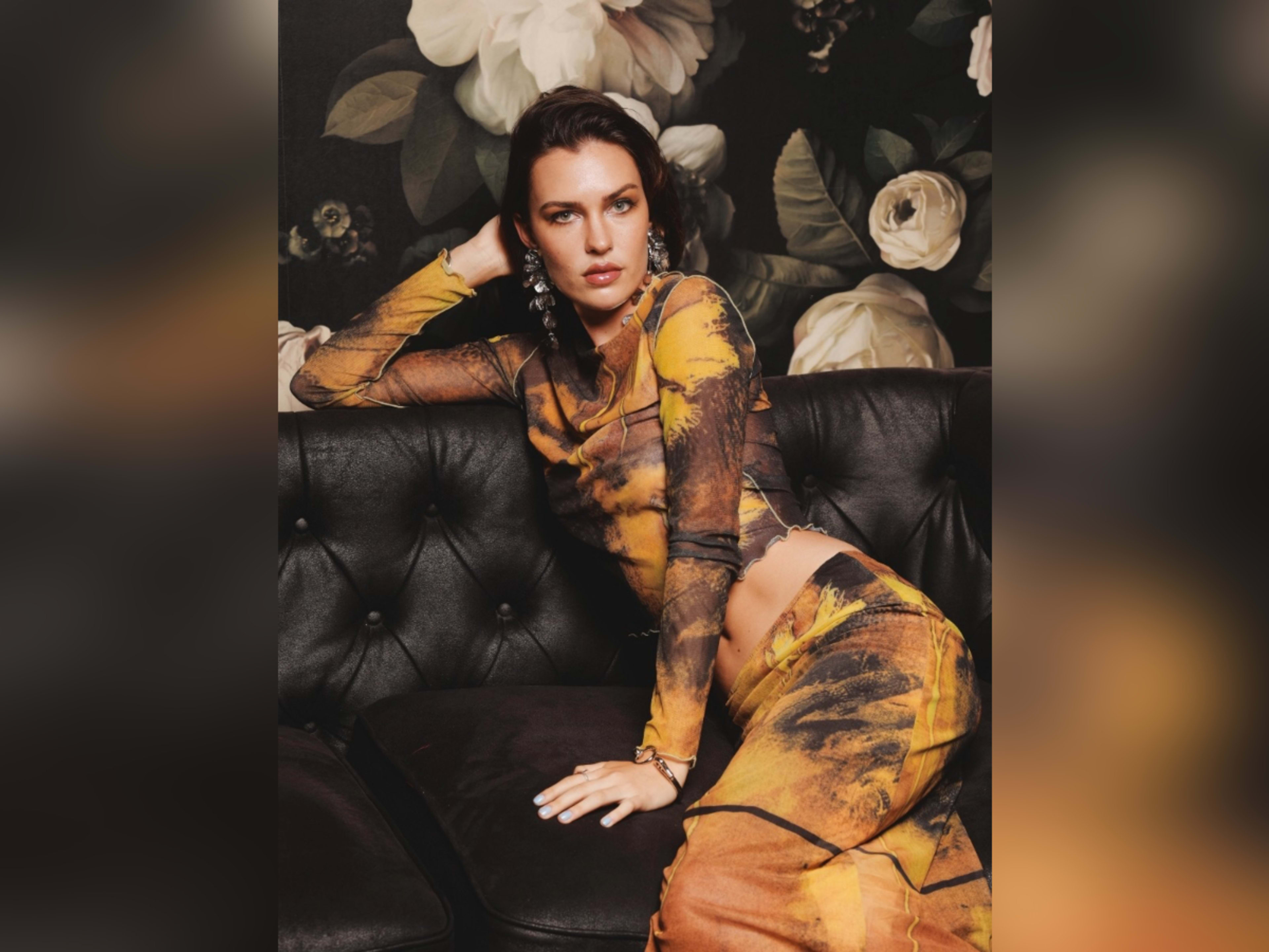 A woman poses on a black leather sofa, wearing an orange and yellow patterned outfit against a floral wallpaper backdrop.