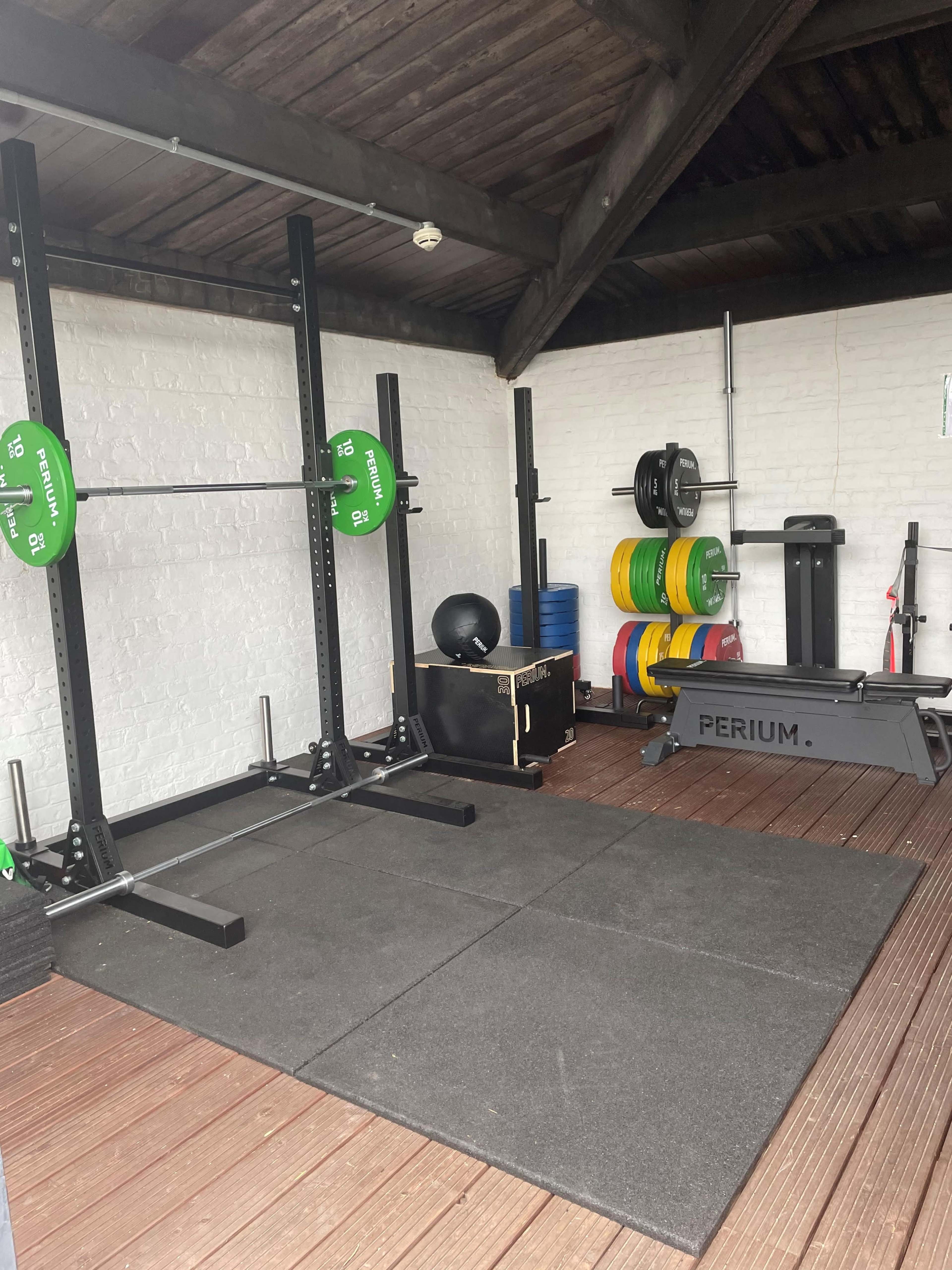 A home gym setup with a squat rack, weights, a rowing machine, and rubber flooring in a wooden structure.