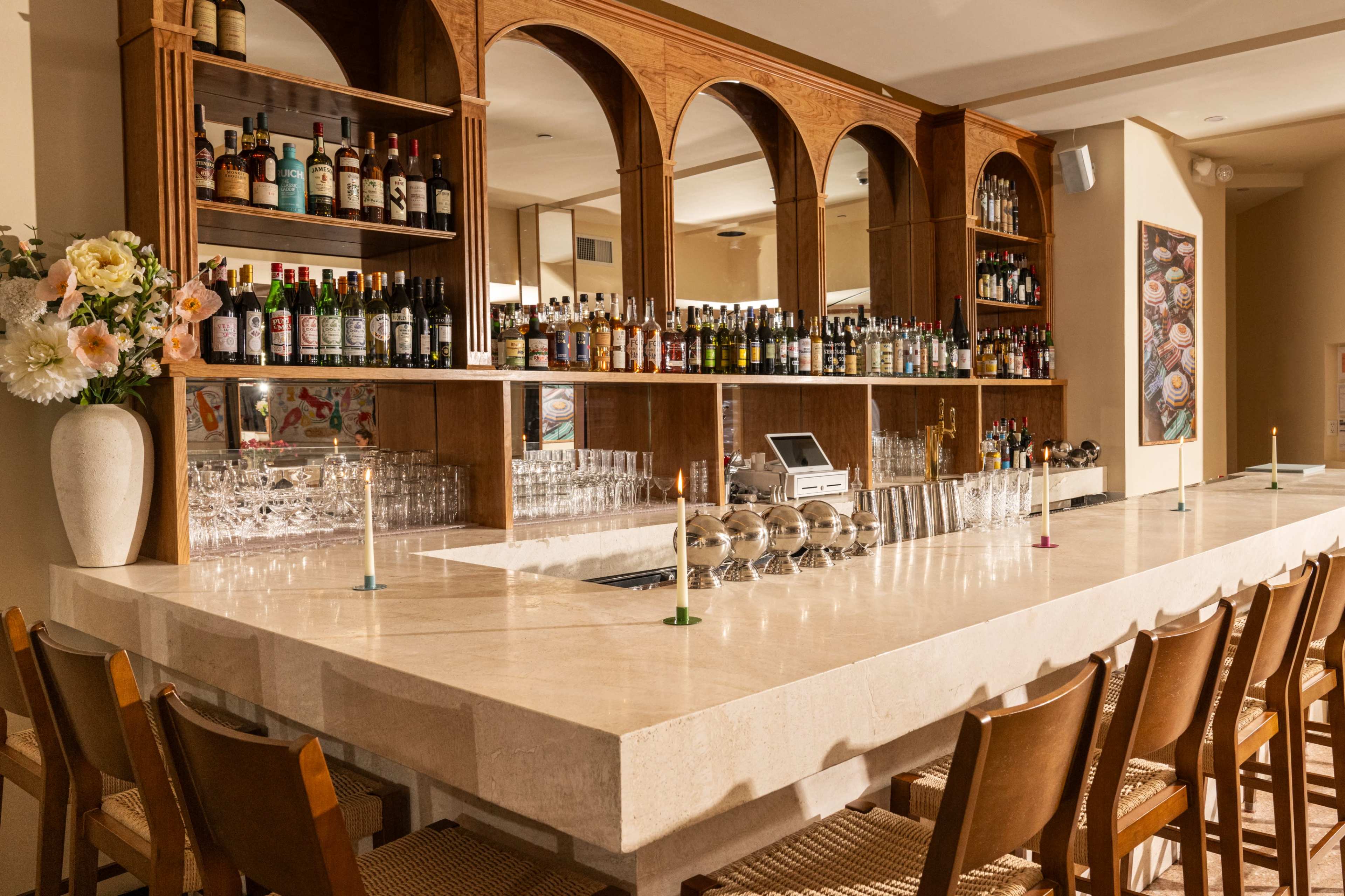 The image shows a modern bar with a polished stone countertop, wooden chairs, and shelves filled with various bottles of liquor and glassware.