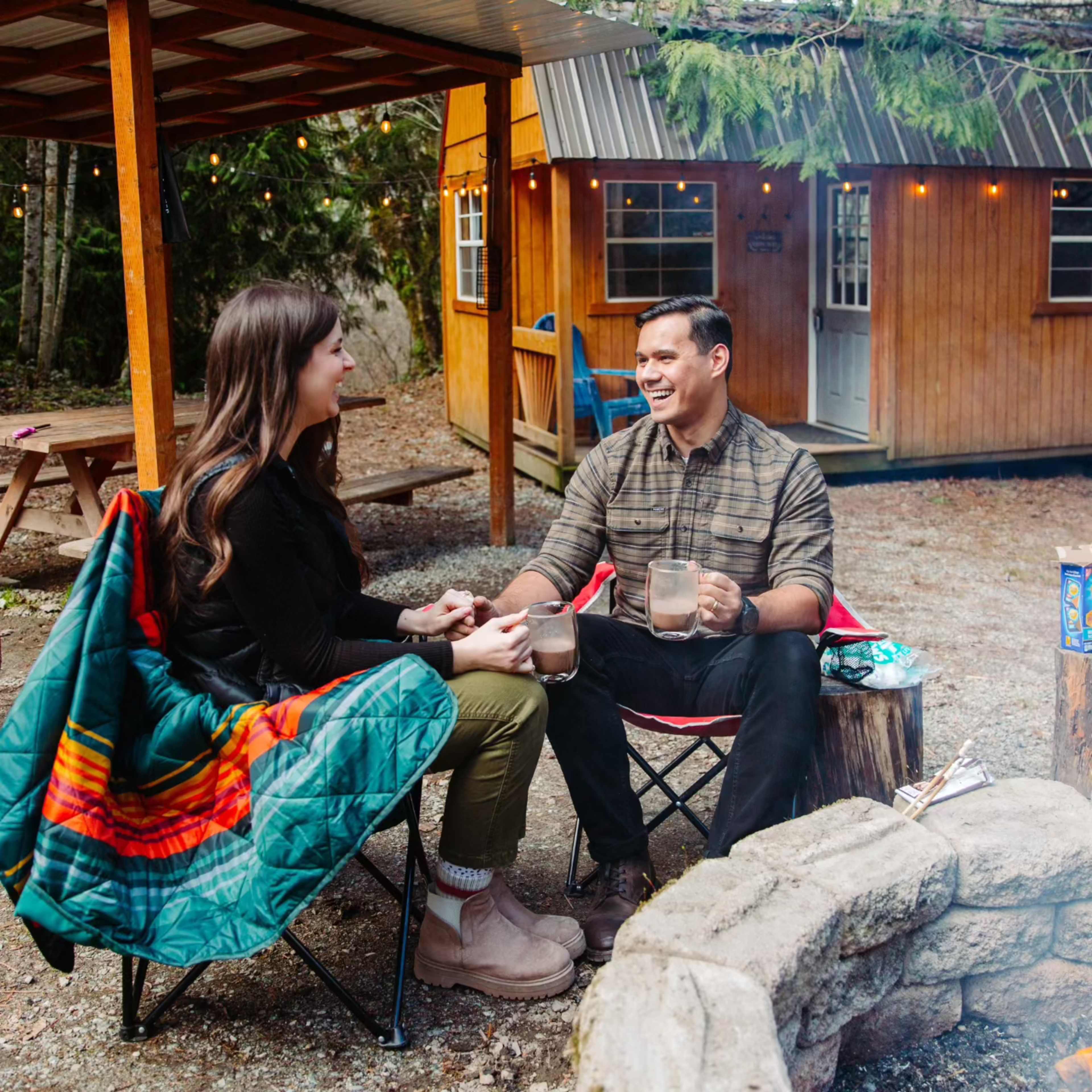 A couple sits in camping chairs outside a cabin, sharing drinks while wrapped in a blanket under string lights.